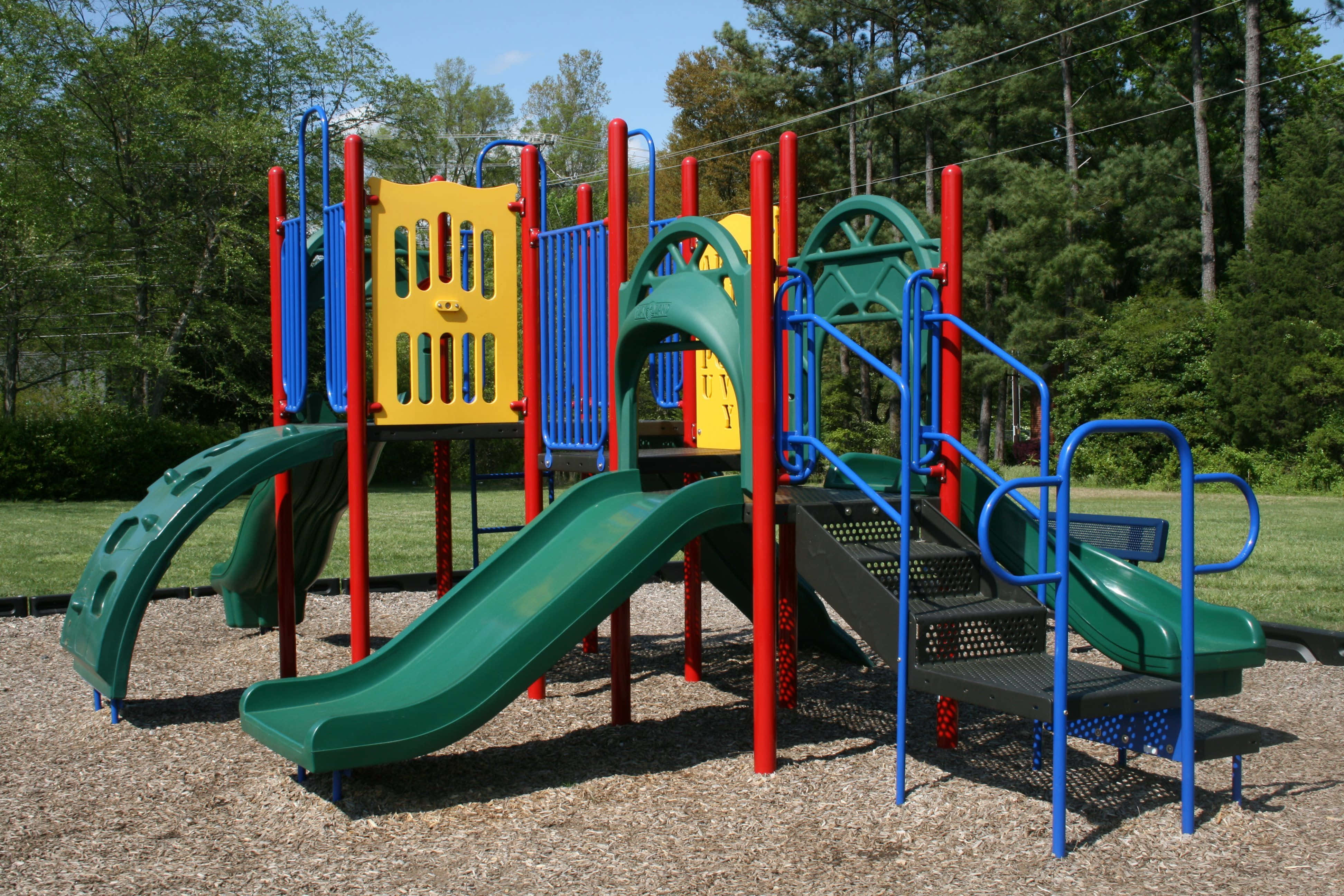 Playground 桌布 壁紙 — 充滿活力的綠色、藍色和黃色遊樂場設備，站在木屑地面上，背後有樹木，在晴朗的藍天下。