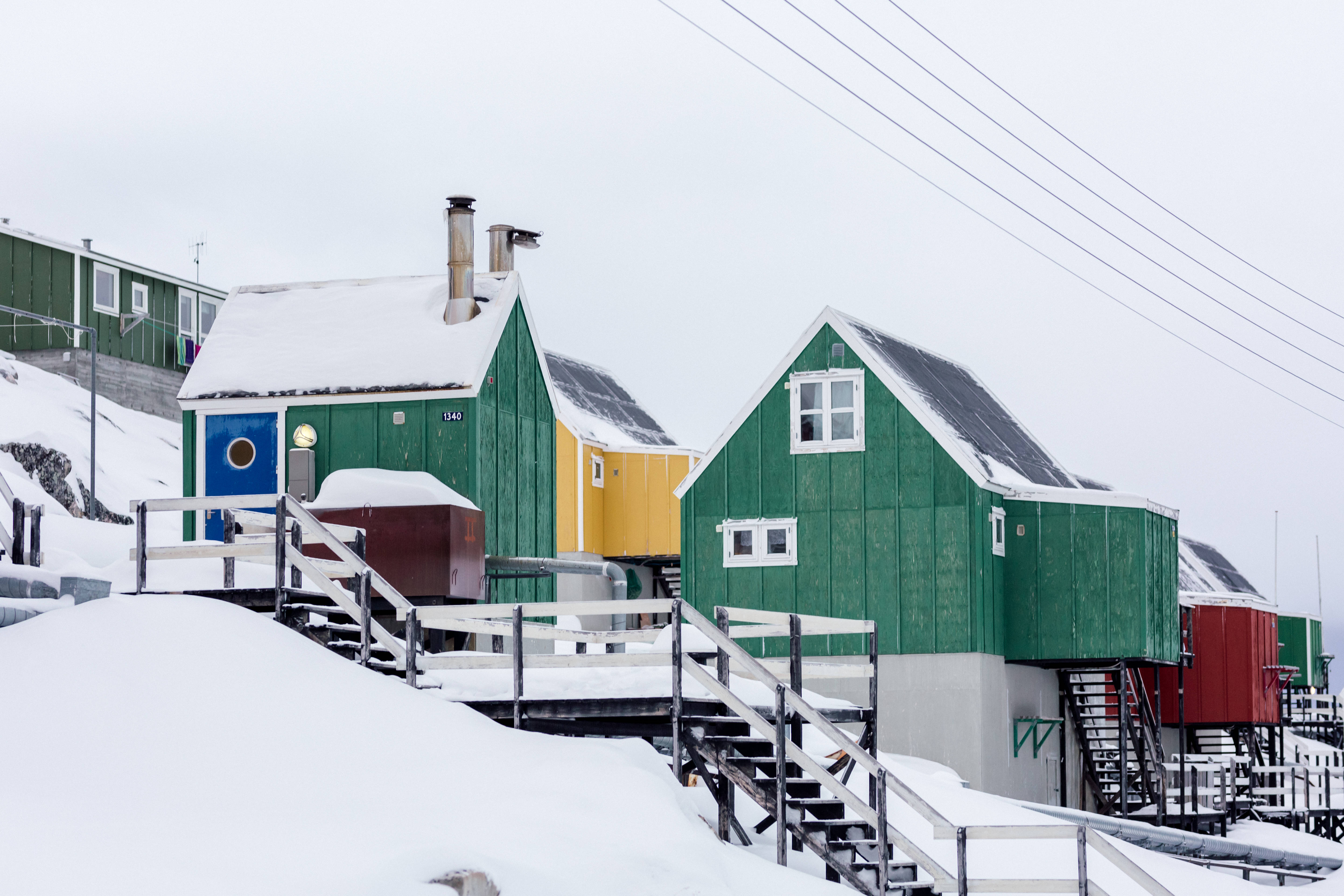 Greenland 桌布 壁紙 — 充滿活力的綠色、黃色和紅色小屋棲息在積雪覆蓋的高蹺上，映襯著灰色的冬日天空。