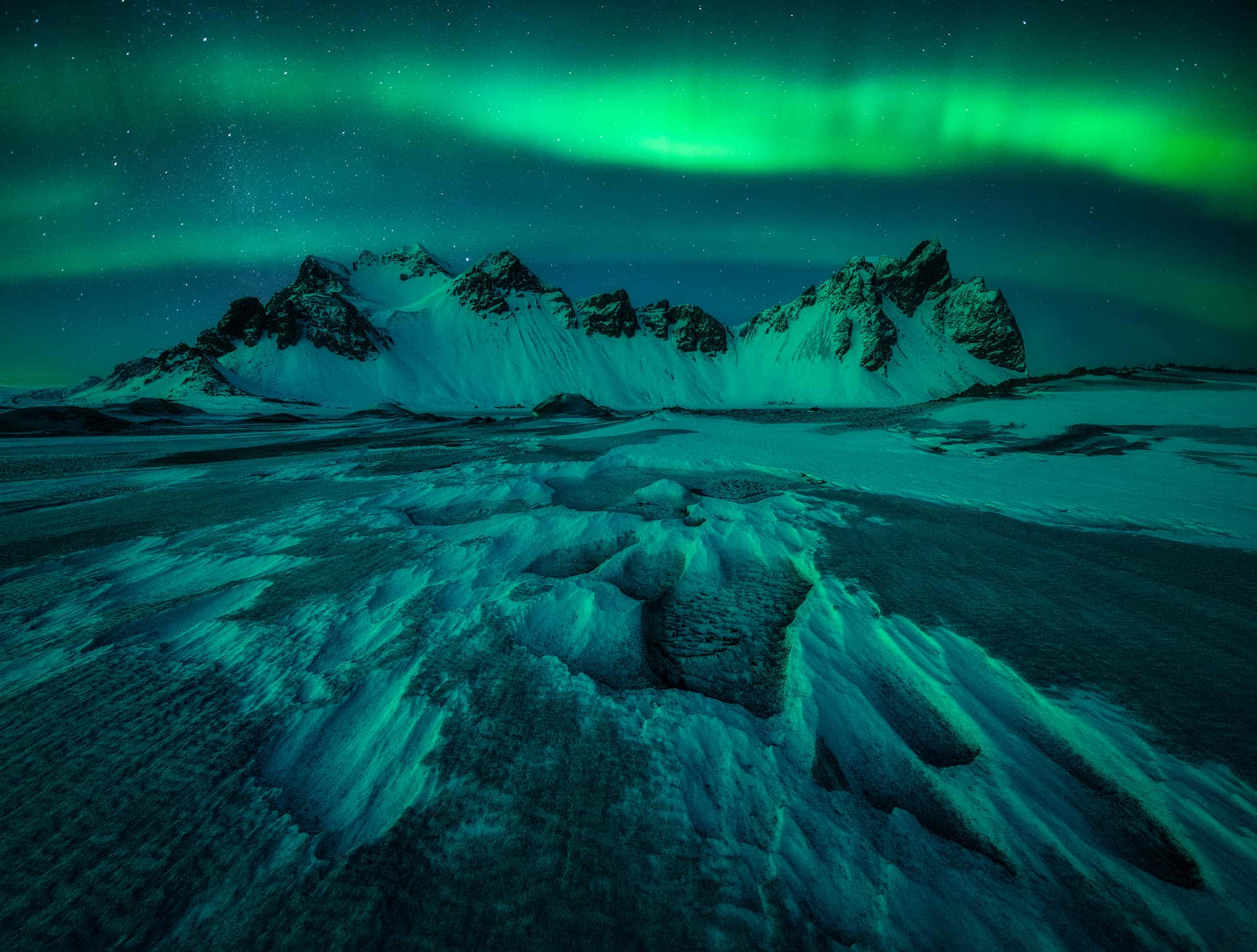 極光 桌布 壁紙 — 充滿活力的綠色北極光在冰雪地形上方的星空夜空下，在積雪的山峰上舞動。