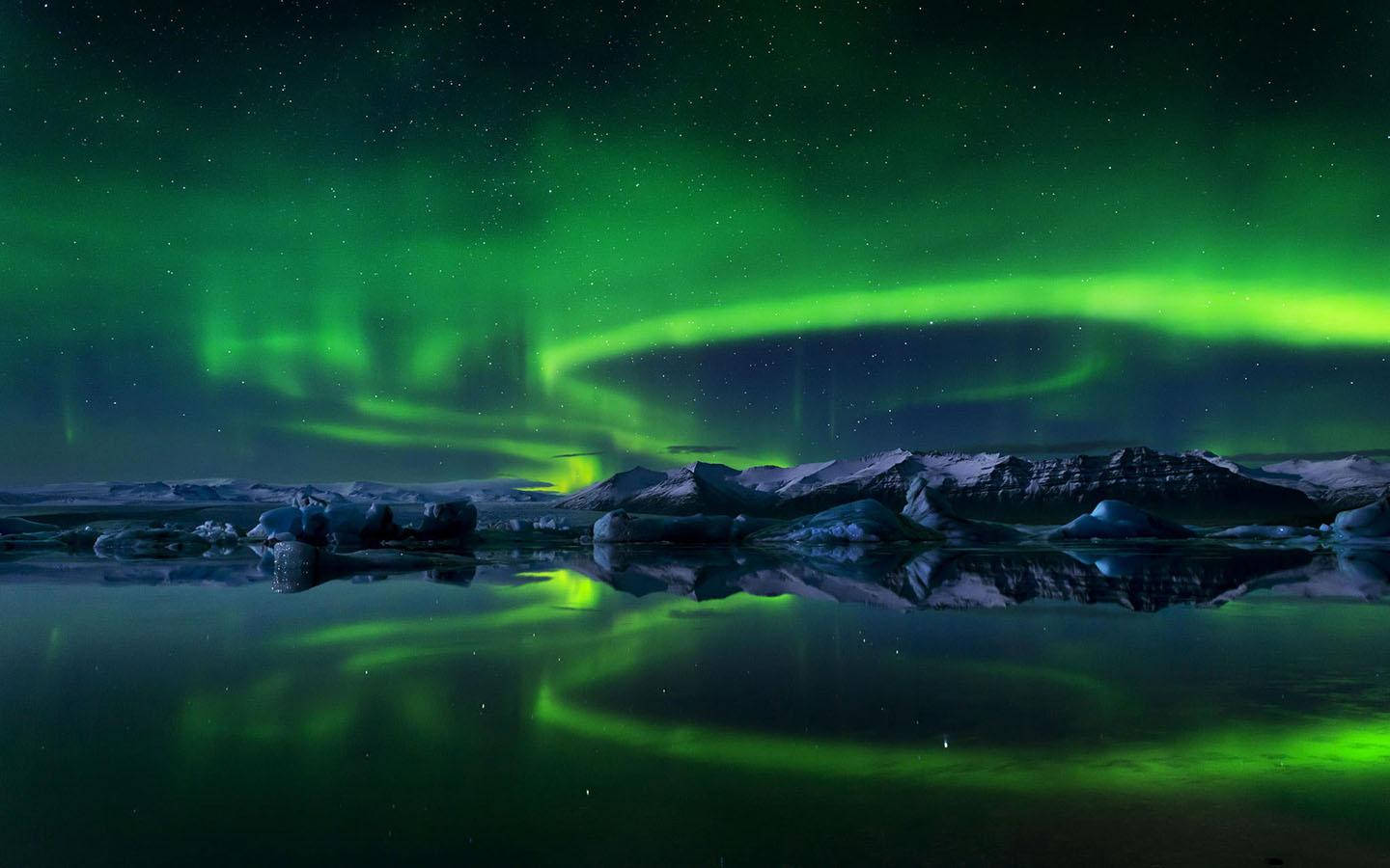 lights 桌布 壁紙 — 充滿活力的綠色北極光在繁星點點的夜空下，在白雪覆蓋的山脈和冰冷的水域上方舞動。