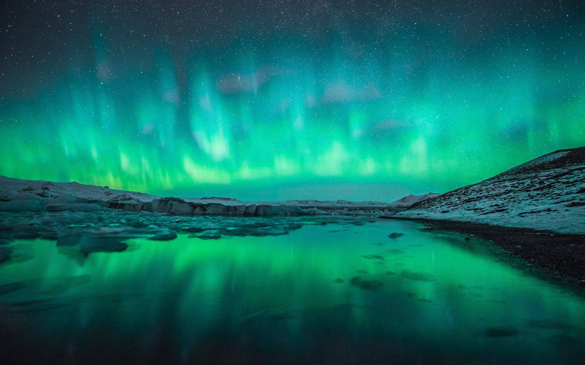 lights 桌布 壁紙 — 充滿活力的綠色北極光在繁星點點的夜空中舞動，倒映在平靜的冰湖中，周圍是白雪覆蓋的山丘。