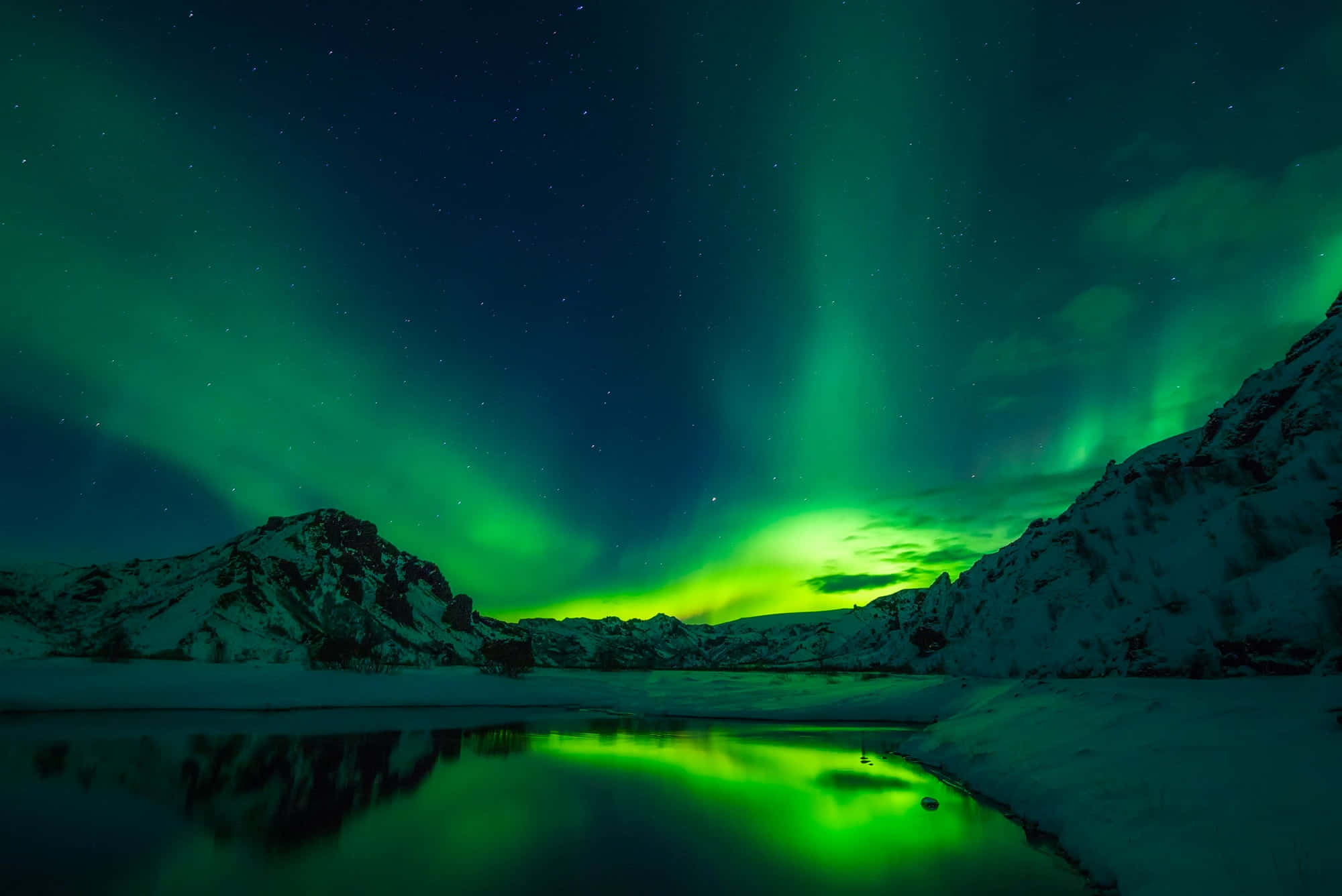 Aurora 桌布 壁紙 — 充滿活力的綠色北極光在繁星點點的夜空中舞動，映照著白雪覆蓋的山脈和反射的冰凍湖泊。