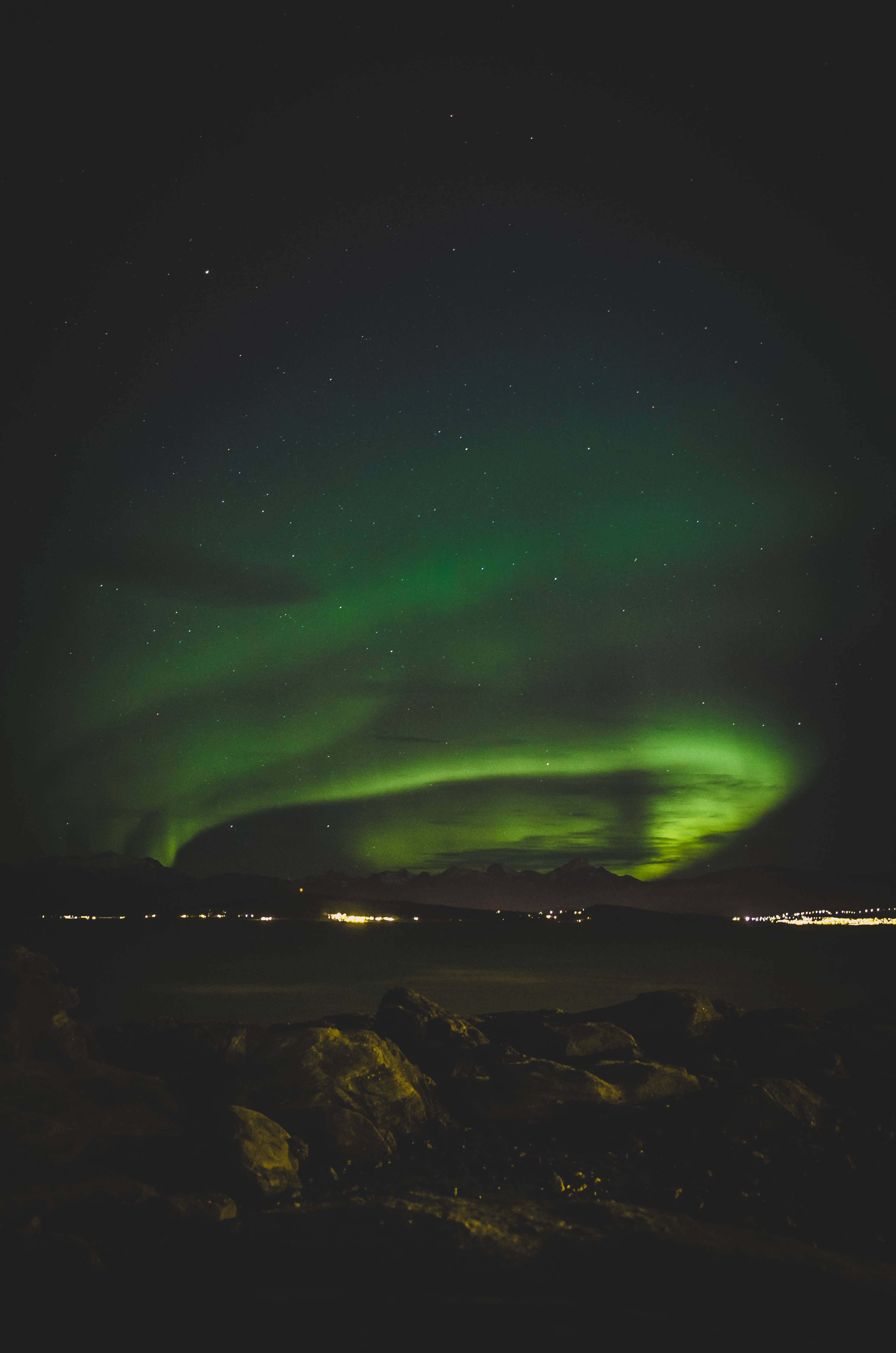 lights 桌布 壁紙 — 充滿活力的綠色北極光在繁星點點的夜空下，在岩石海岸線和遠處城市燈光上方舞動。