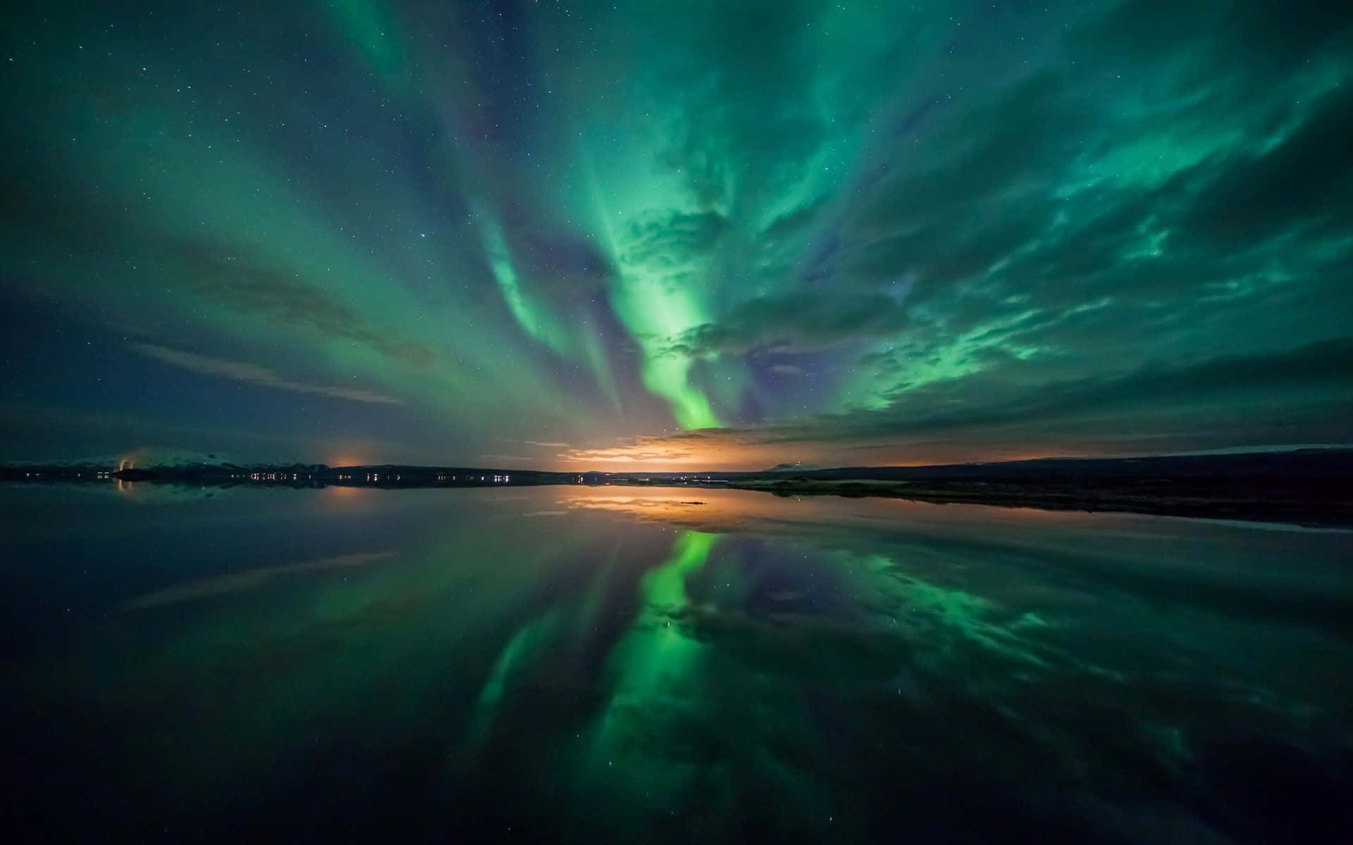 Aurora 桌布 壁紙 — 充滿活力的綠色北極光在繁星點點的夜空中舞動，倒映在平靜的湖面上，地平線上有遠處的燈光。