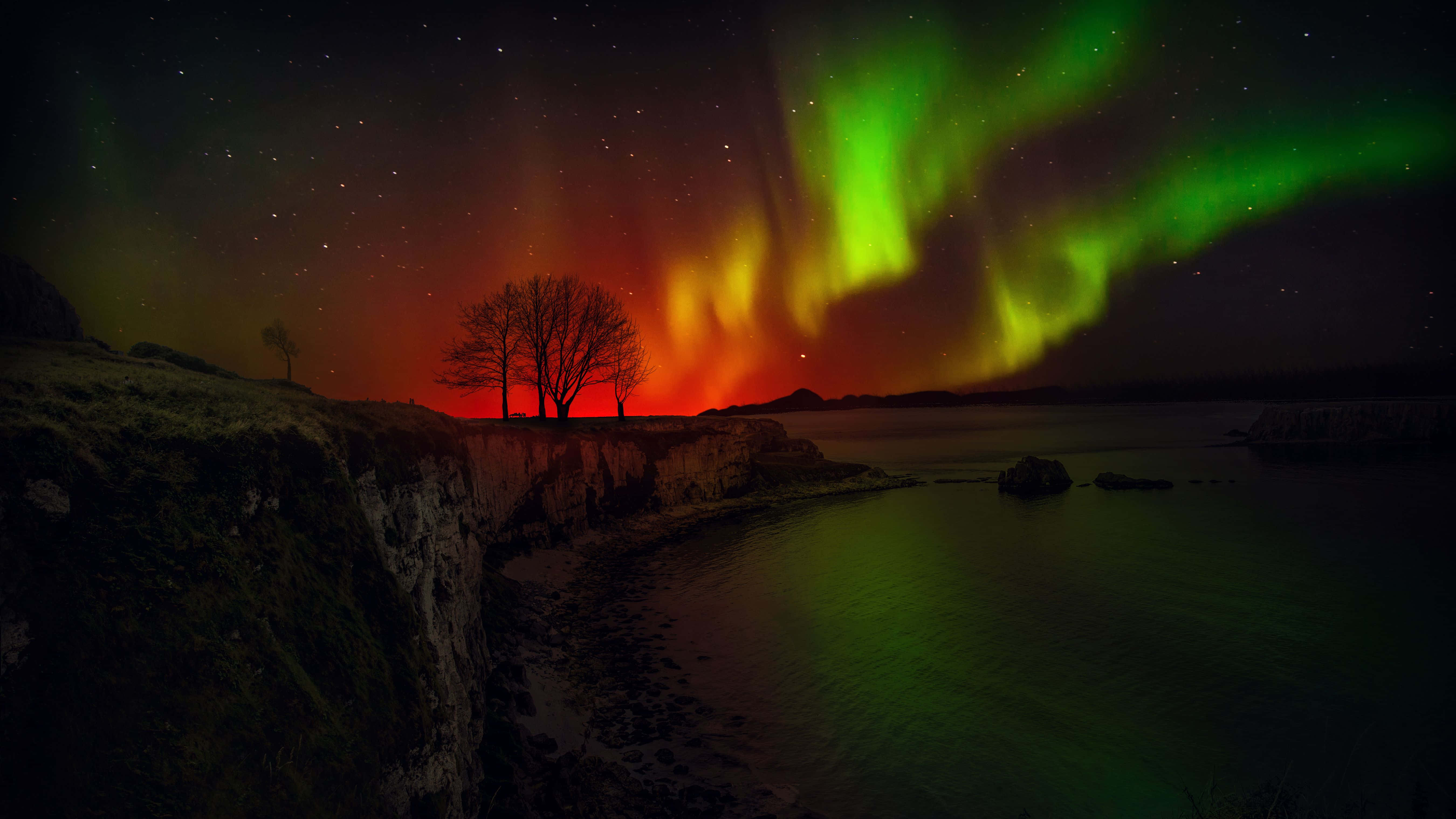 Aurora 桌布 壁紙 — 充滿活力的綠色和橙色北極光在星空下舞動，懸崖海岸場景中有剪影樹木。