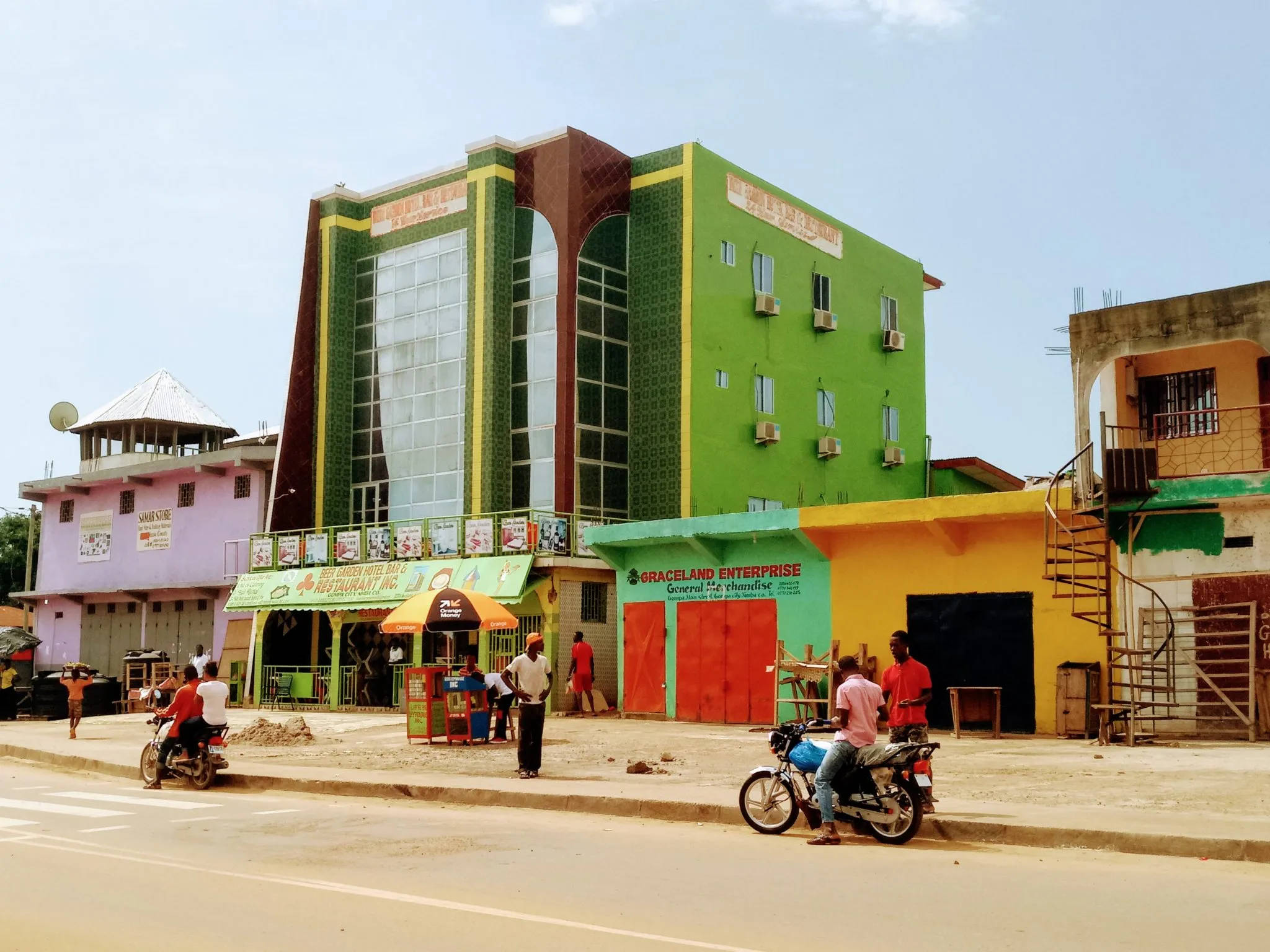 Liberia 桌布 壁紙 — 充滿活力的綠色和黃色玻璃帷幕建築，在繁忙的街道場景中高聳，摩托車和行人穿梭於晴朗的藍天下。