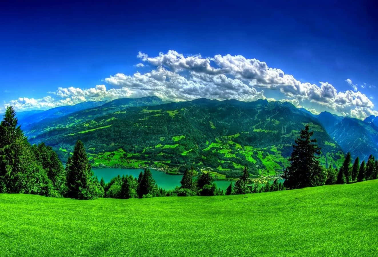 Lake 桌布 壁紙 — 充滿活力的綠色草甸向下傾斜到一個碧綠色的湖泊，湖泊坐落在森林覆蓋的山脈之間，上方是明亮的藍天和蓬鬆的白雲。