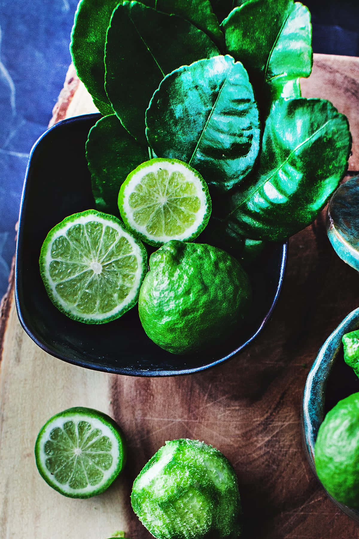 citrus 桌布 壁紙 — 充滿活力的綠色萊姆和葉子放在深色碗中，置於質樸的木質表面上，背景為清涼的藍色。