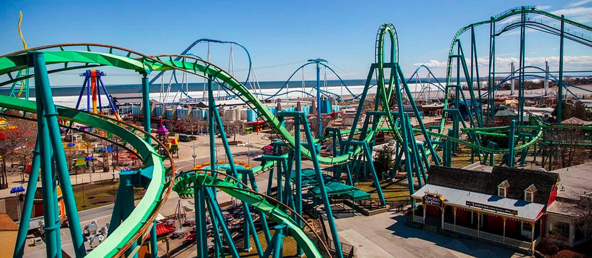 Roller Coaster 桌布 壁紙 — 充滿活力的綠色雲霄飛車在俯瞰海濱遊樂園的清澈藍天下蜿蜒盤旋。