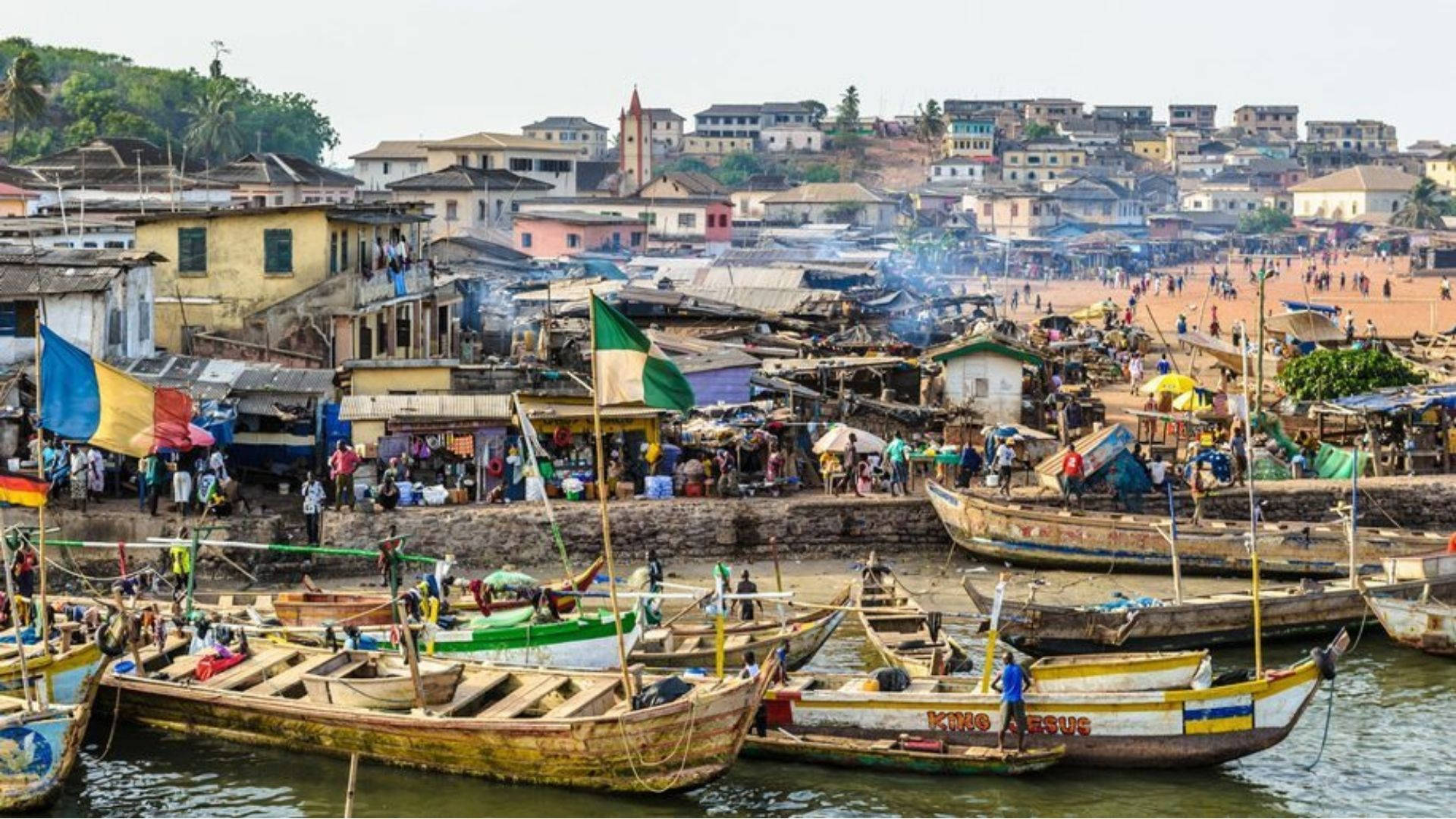 Ghana 桌布 壁紙 — 充滿活力的繁忙港口，色彩繽紛的木船停泊在密集的沿海建築前，在明亮的日光下。