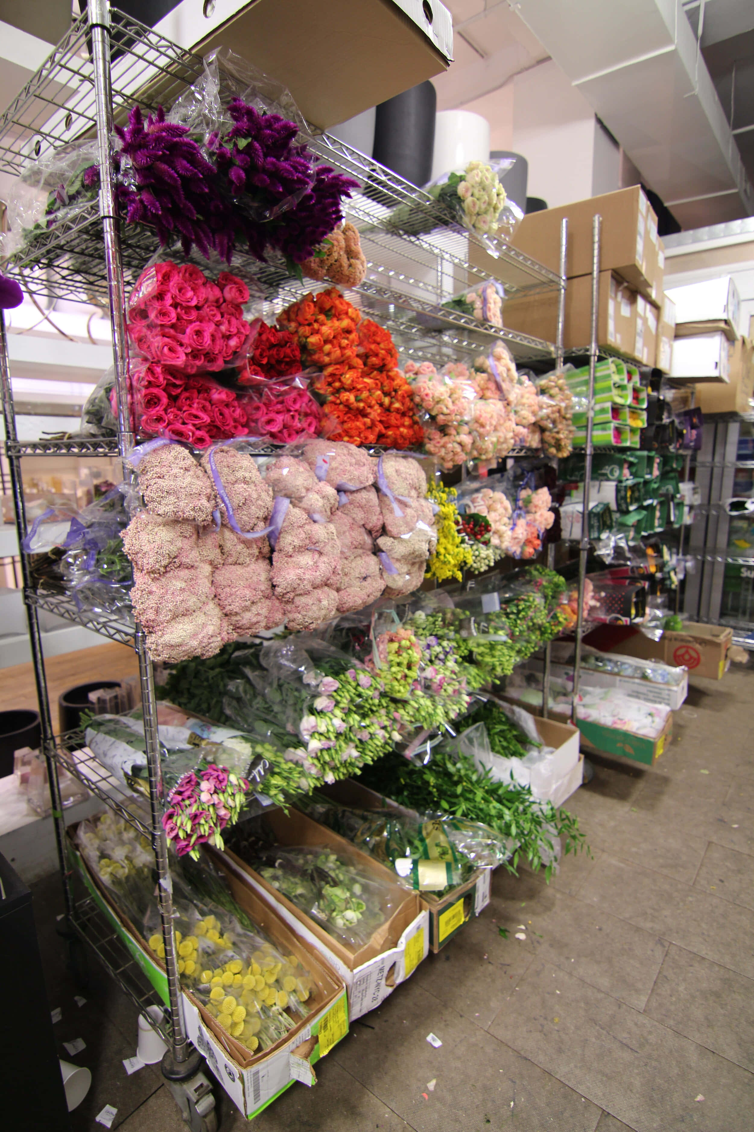 Flower Market 桌布 壁紙 — 充滿活力的花店通道展示色彩繽紛的花束和莖部，用塑膠包裝包裹在金屬架上，置於明亮的螢光燈下。