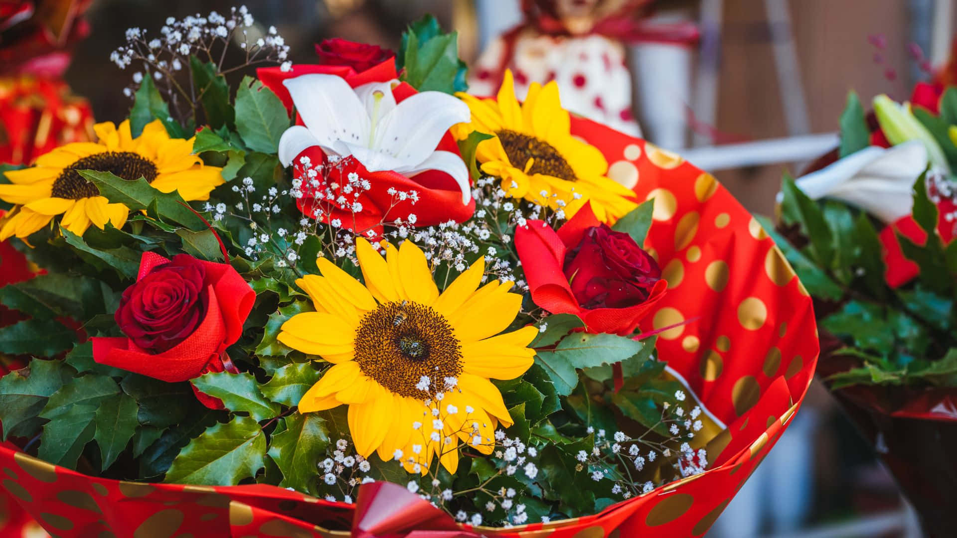 Roses 桌布 壁紙 — 充滿活力的花束，包含向日葵、紅玫瑰和白百合，用帶有金色圓點的節慶紅紙包裹。
