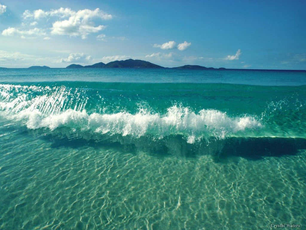 海洋 桌布 壁紙 — 充滿活力的藍綠色波浪帶著白色泡沫衝擊清澈的淺水，露出沙質底部，上方是明亮的藍天和散落的雲朵。