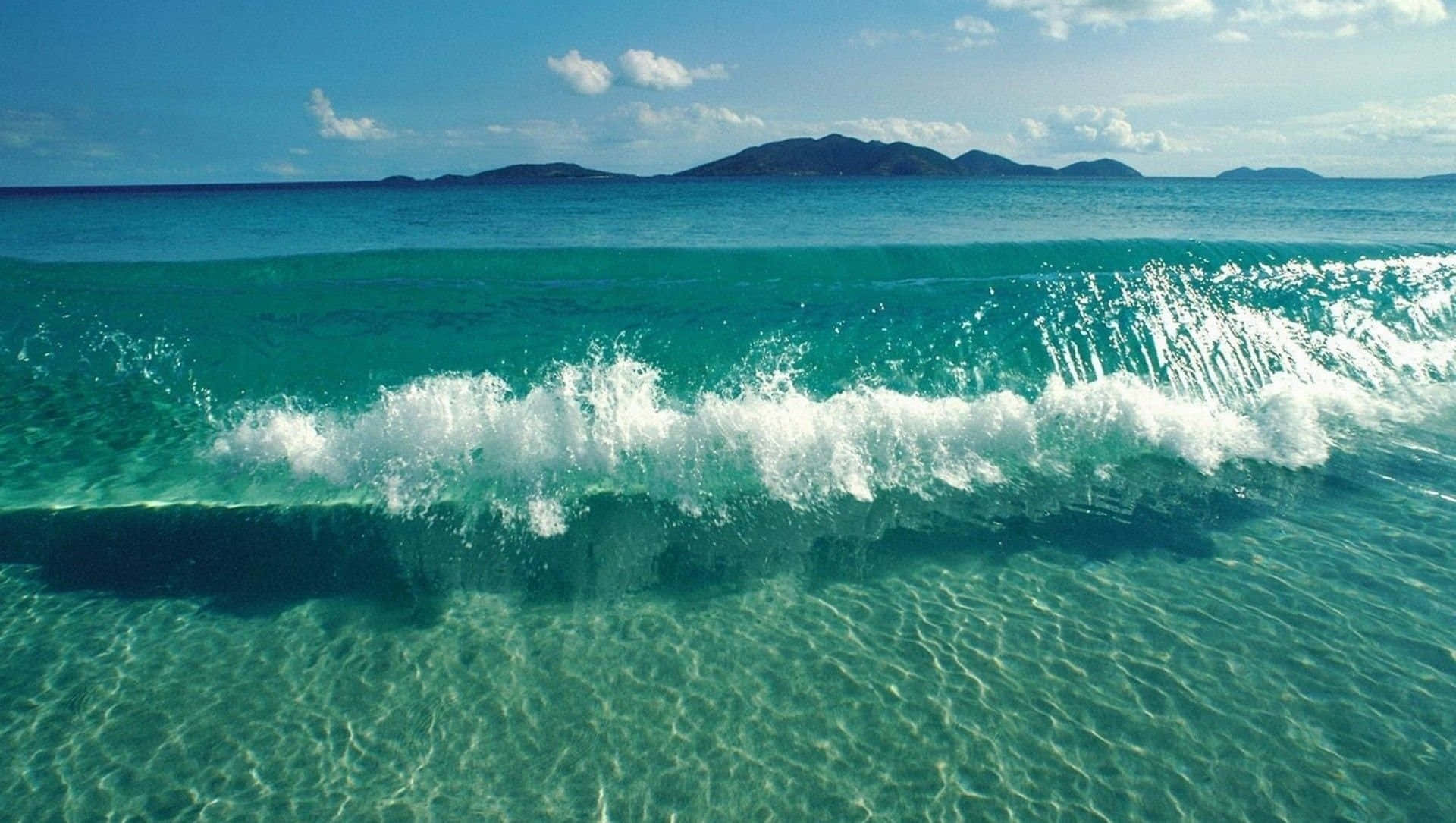 waves 桌布 壁紙 — 充滿活力的藍綠色波浪帶著白色泡沫拍打清澈的淺水，天空湛藍，遠處有島嶼。