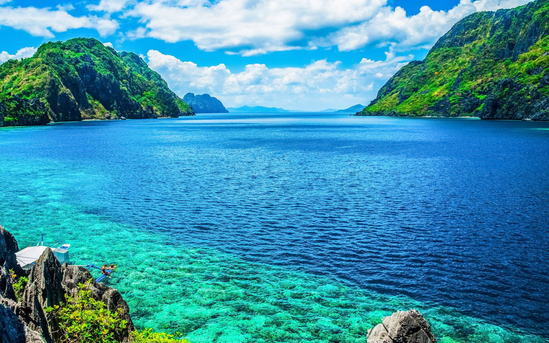Island 桌布 壁紙 — 充滿活力的藍綠色海水 nestled 在鬱鬱蔥蔥的綠色懸崖之間，在明亮的藍天下有蓬鬆的白雲。