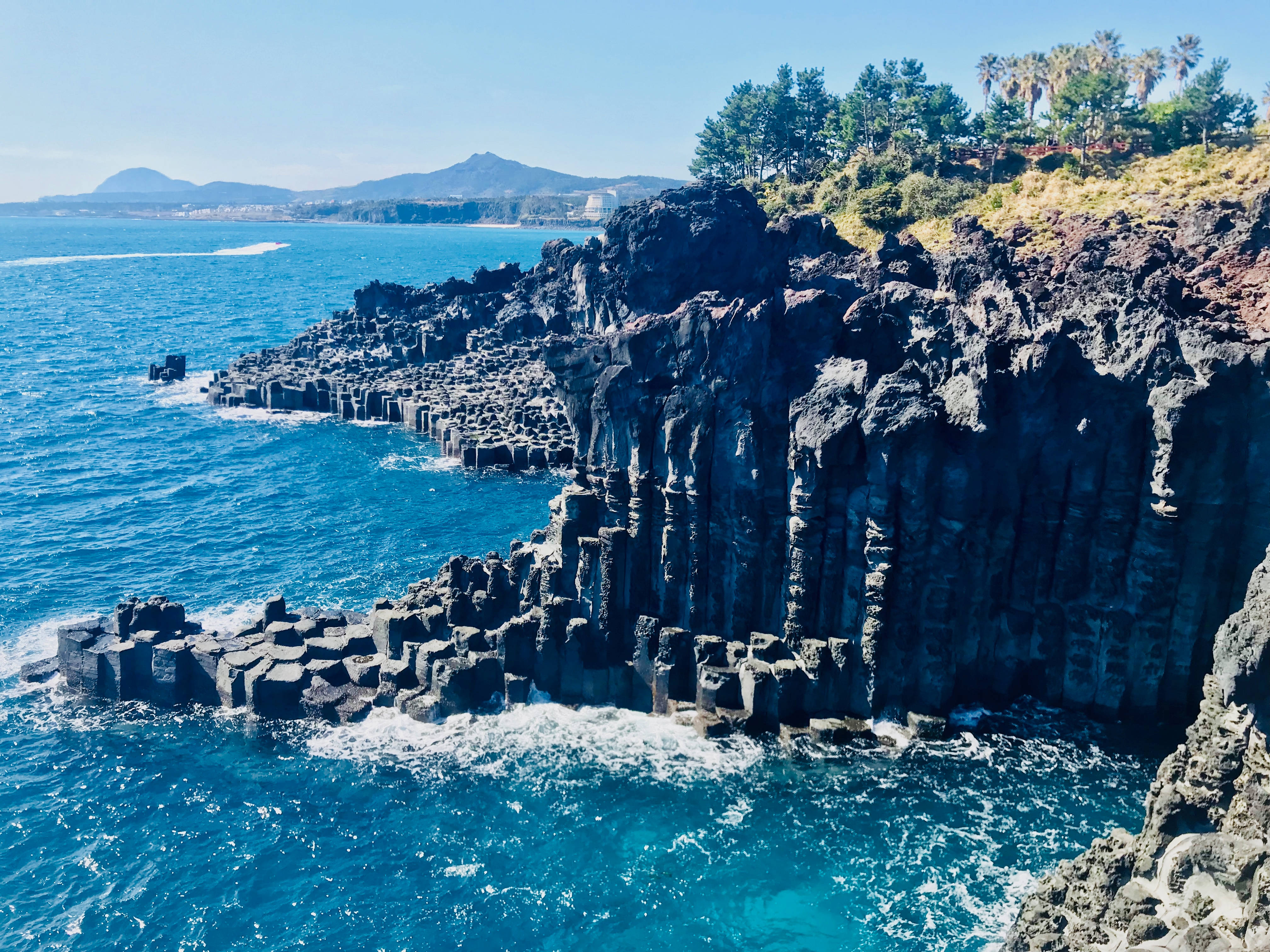 Jeju 桌布 壁紙 — 充滿活力的藍綠色海洋在清澈的藍天下衝擊著戲劇性的黑色玄武岩柱。