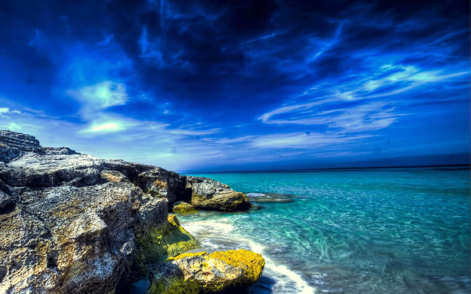 Beach Desktop 桌布 壁紙 — 充滿活力的藍綠色海浪拍打崎嶇的懸崖，背景是帶有縷縷白雲的戲劇性深藍色天空。