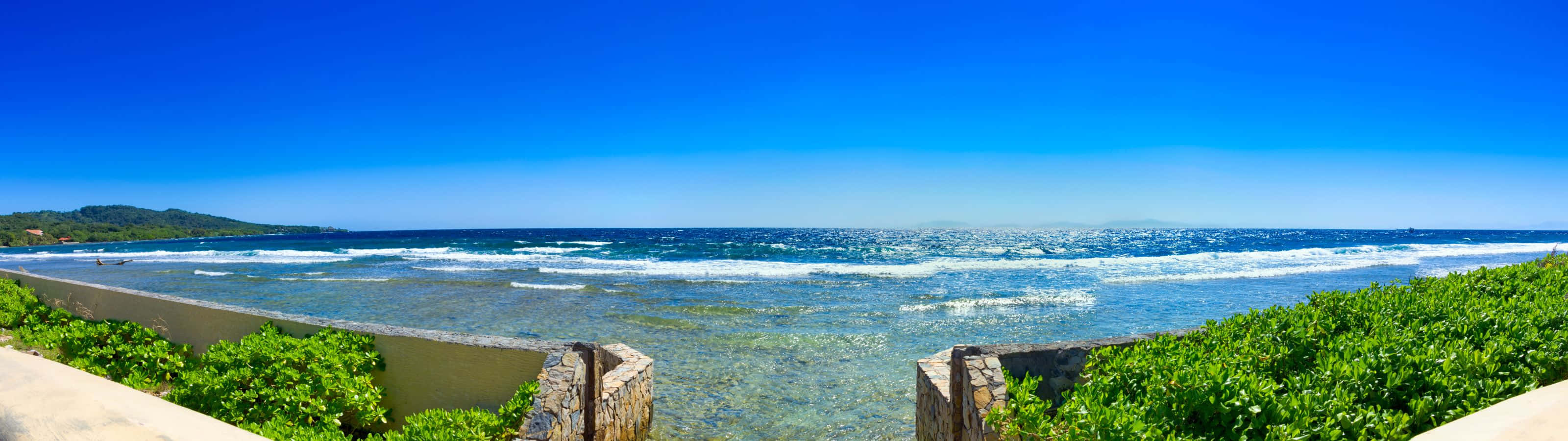 3200x900 桌布 壁紙 — 充滿活力的藍綠色海浪拍打著石牆，石牆周圍環繞著茂密的綠色樹葉，在清澈的藍天下。