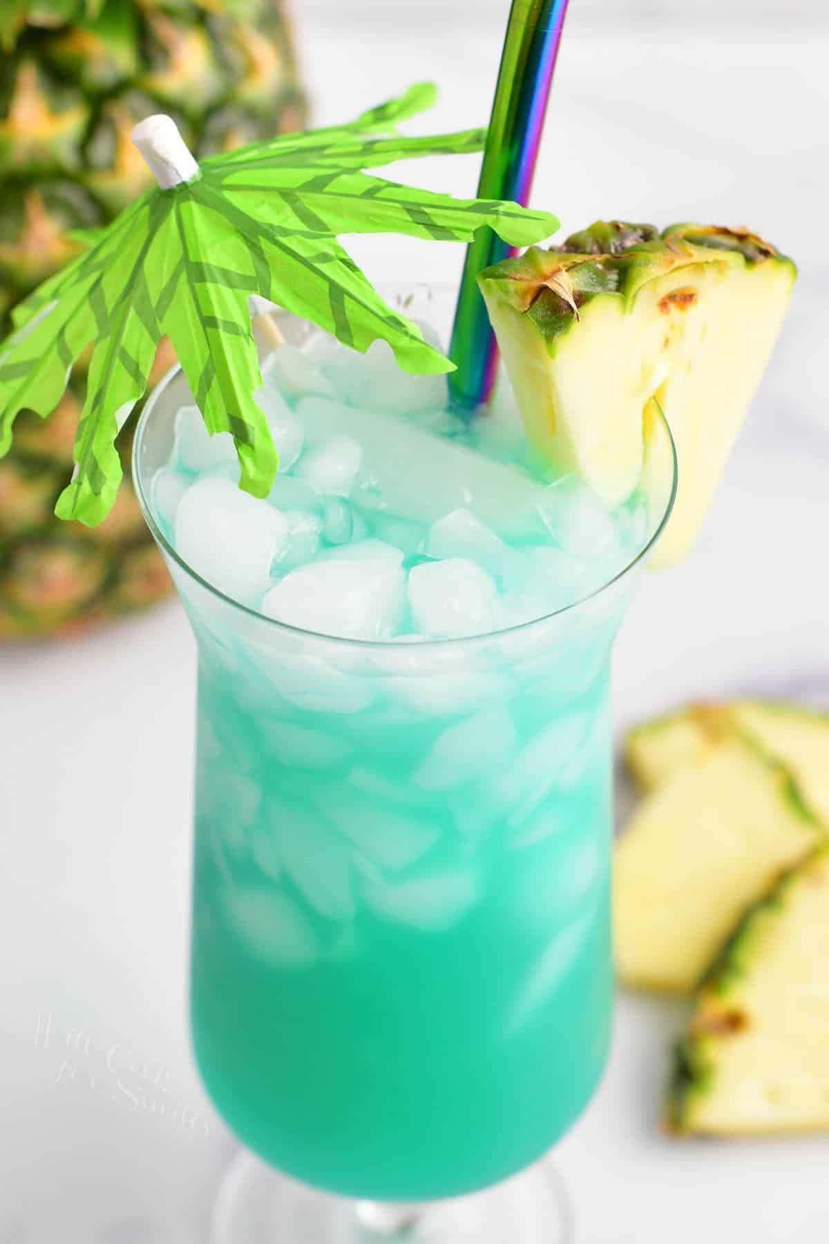 Blue Hawaii 桌布 壁紙 — 充滿活力的藍綠色熱帶飲料，裝滿冰塊，並以鳳梨片和綠色紙傘裝飾。