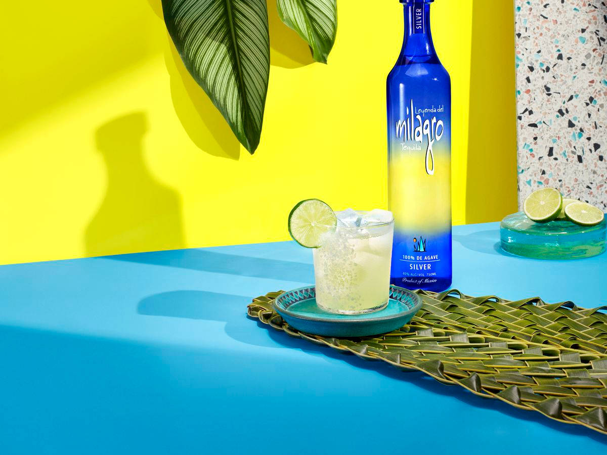 margaritas 桌布 壁紙 — 充滿活力的藍色Milagro Silver龍舌蘭酒瓶和以萊姆裝飾的雞尾酒，放在編織墊上，背景是鮮黃色的牆壁，熱帶樹葉投下陰影。