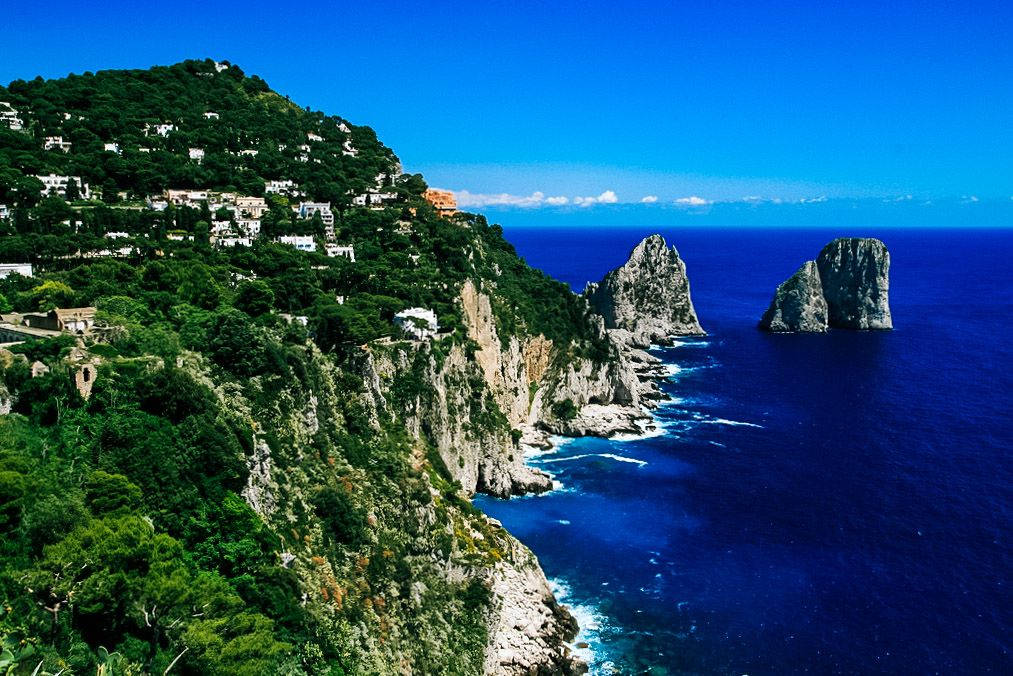 Capri 桌布 壁紙 — 充滿活力的藍色海洋拍打著陡峭的懸崖，懸崖上點綴著白色的房屋和茂密的綠色植物，天空晴朗。