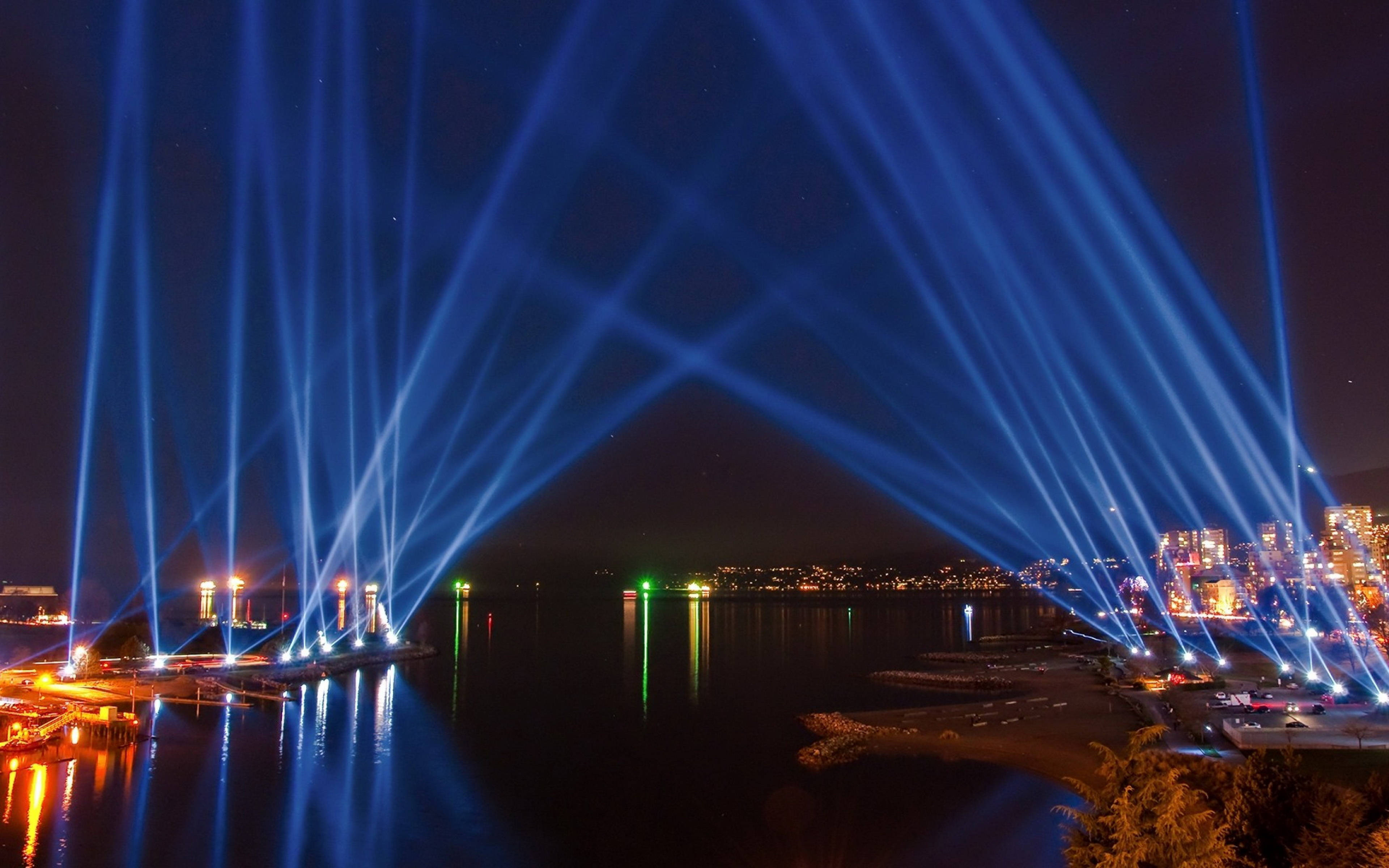 lights 桌布 壁紙 — 充滿活力的藍色激光束在夜間穿過黑暗的海濱，照亮水面和城市天際線。