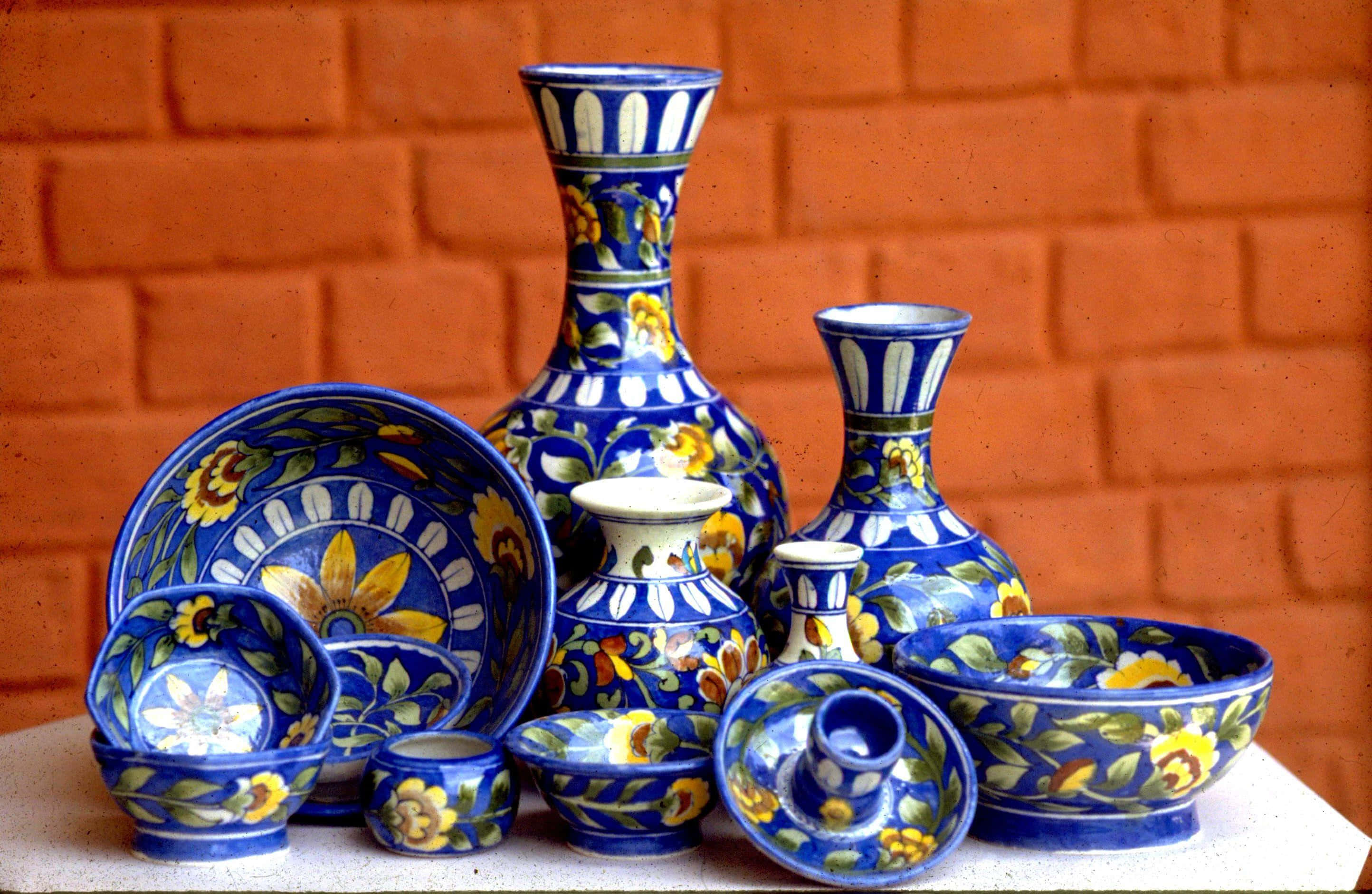 Ceramics 桌布 壁紙 — 充滿活力的藍色陶瓷碗和花瓶，帶有黃色花卉圖案，排列在白色表面上，背景是溫暖的橙色磚牆。