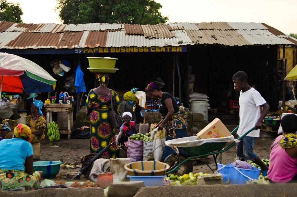 Liberia 桌布 壁紙 — 充滿活力的街市場景，身穿彩色圖案裙子的女性在生鏽的波紋鐵皮屋頂下平衡貨物並出售新鮮農產品。