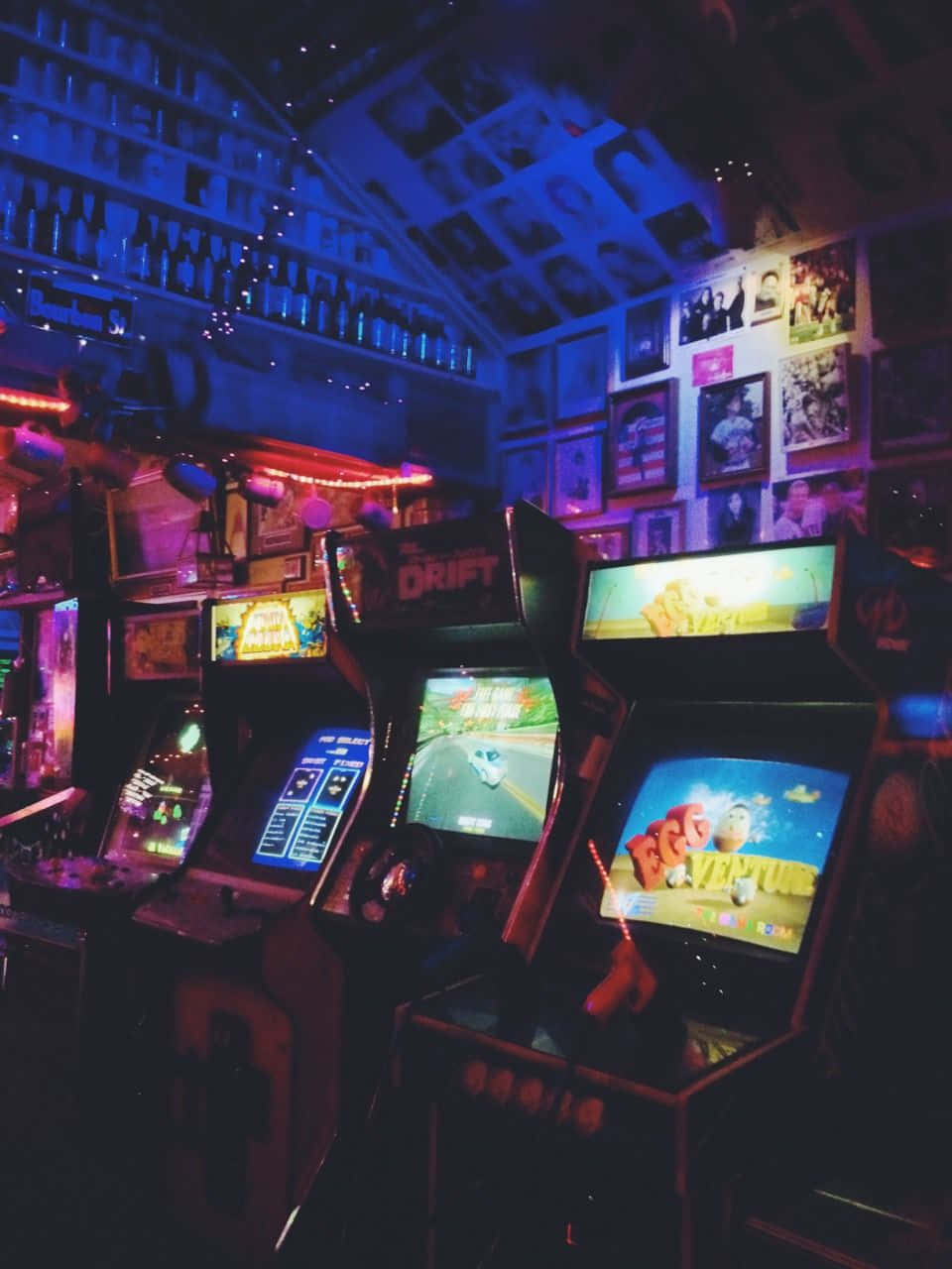 Arcade Aesthetic 桌布 壁紙 — 充滿活力的街機在霓虹燈下閃耀，位於一個光線昏暗的復古遊戲室，裝飾著海報和酒瓶架。