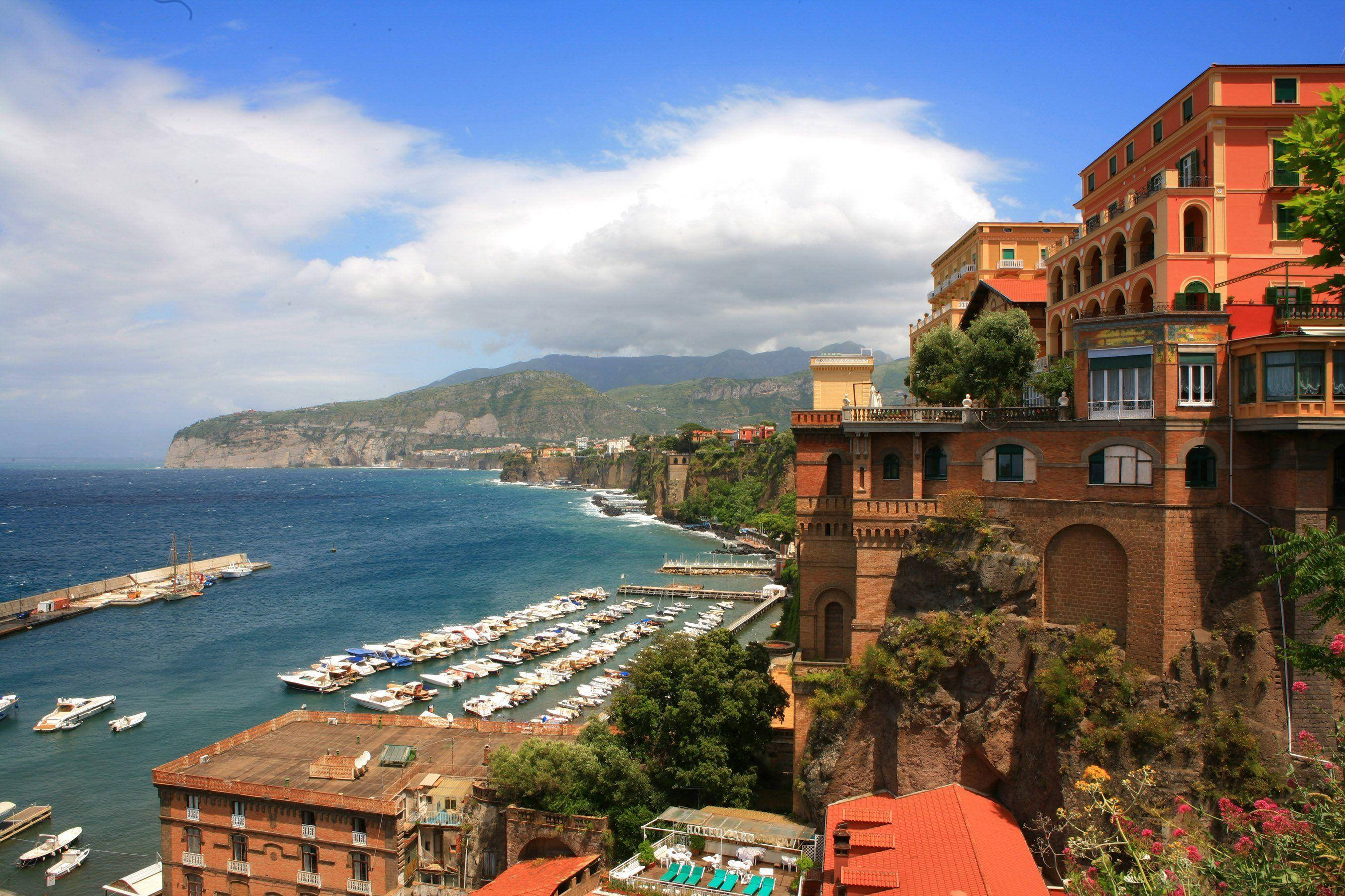 Naples 桌布 壁紙 — 充滿活力的赤陶建築依附在岩石懸崖上，俯瞰著停滿白色船隻的碼頭，上方是明亮的藍天。