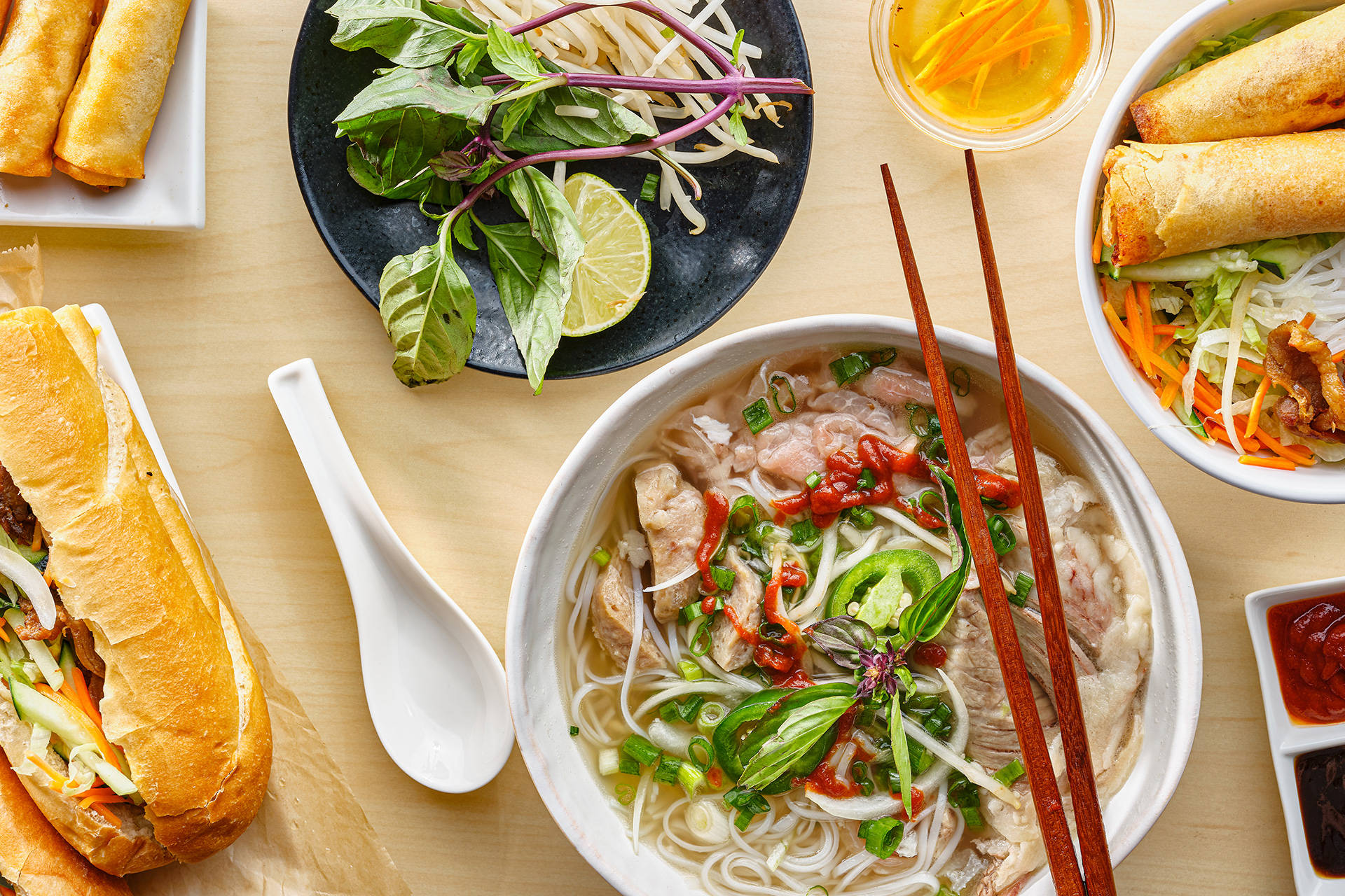 Food Photography 桌布 壁紙 — 充滿活力的越南菜餚，包括河粉、春捲和越南法包三明治，擺放在淺色木桌上。