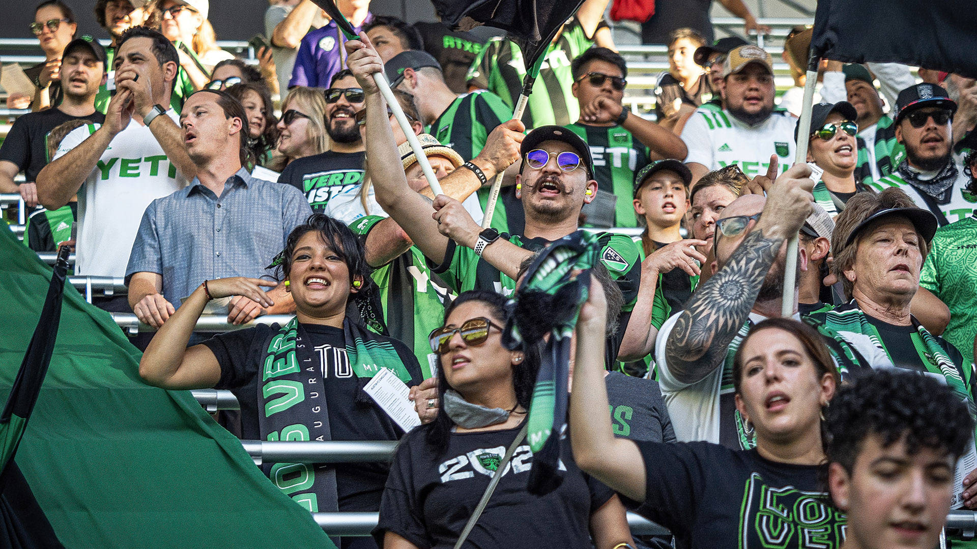 Austin Fc 桌布 壁紙 — 充滿活力的足球迷穿著綠色和黑色球衣，在體育場看台上揮舞旗幟和圍巾，熱情地歡呼。