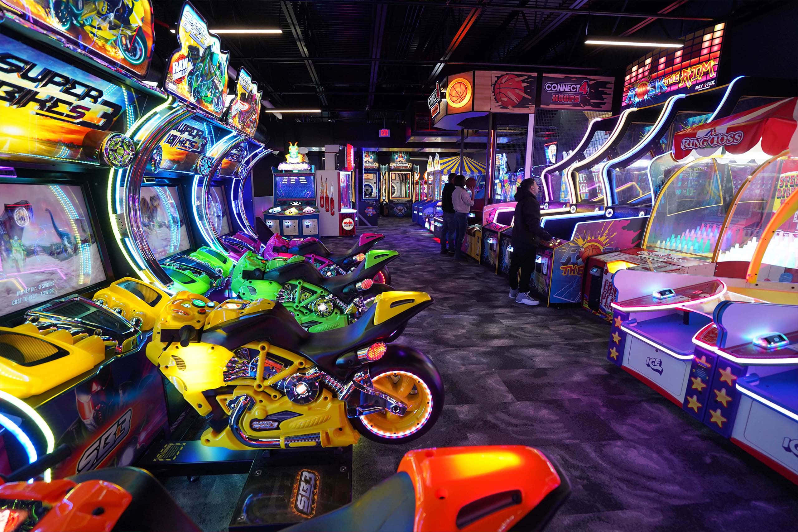 Arcade Games 桌布 壁紙 — 充滿活力的遊戲機室，內有發光的賽車遊戲機和霓虹燈，在黑暗的地板上反射。