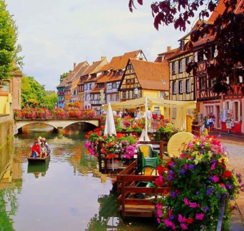 France 桌布 壁紙 — 充滿活力的運河場景，色彩繽紛的半木結構房屋、盛開的花朵，以及一艘載著遊客的小船在明亮的天空下。