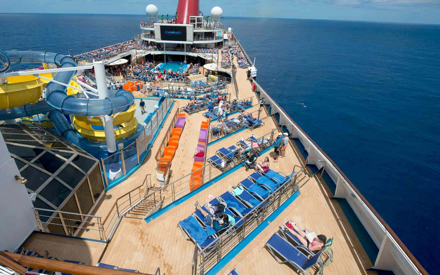 Carnival Conquest 桌布 壁紙 — 充滿活力的郵輪甲板，乘客在藍色椅子上放鬆，旁邊是色彩繽紛的水滑梯，背景是深藍色的海洋。