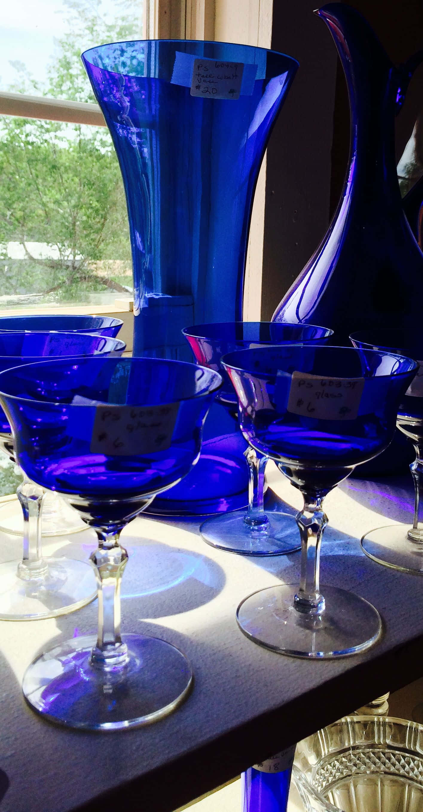 Cobalt Blue 桌布 壁紙 — 充滿活力的鈷藍色玻璃器皿在沐浴著自然日光的窗台上閃閃發光，外面可見綠色植物。