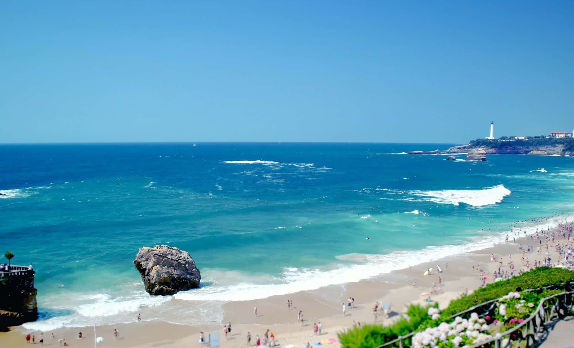 Summer Beach 桌布 壁紙 — 充滿活力的陽光沙灘場景，碧綠的海浪拍打沙灘，沙灘上散佈著遊客和一個突出的岩石形成。