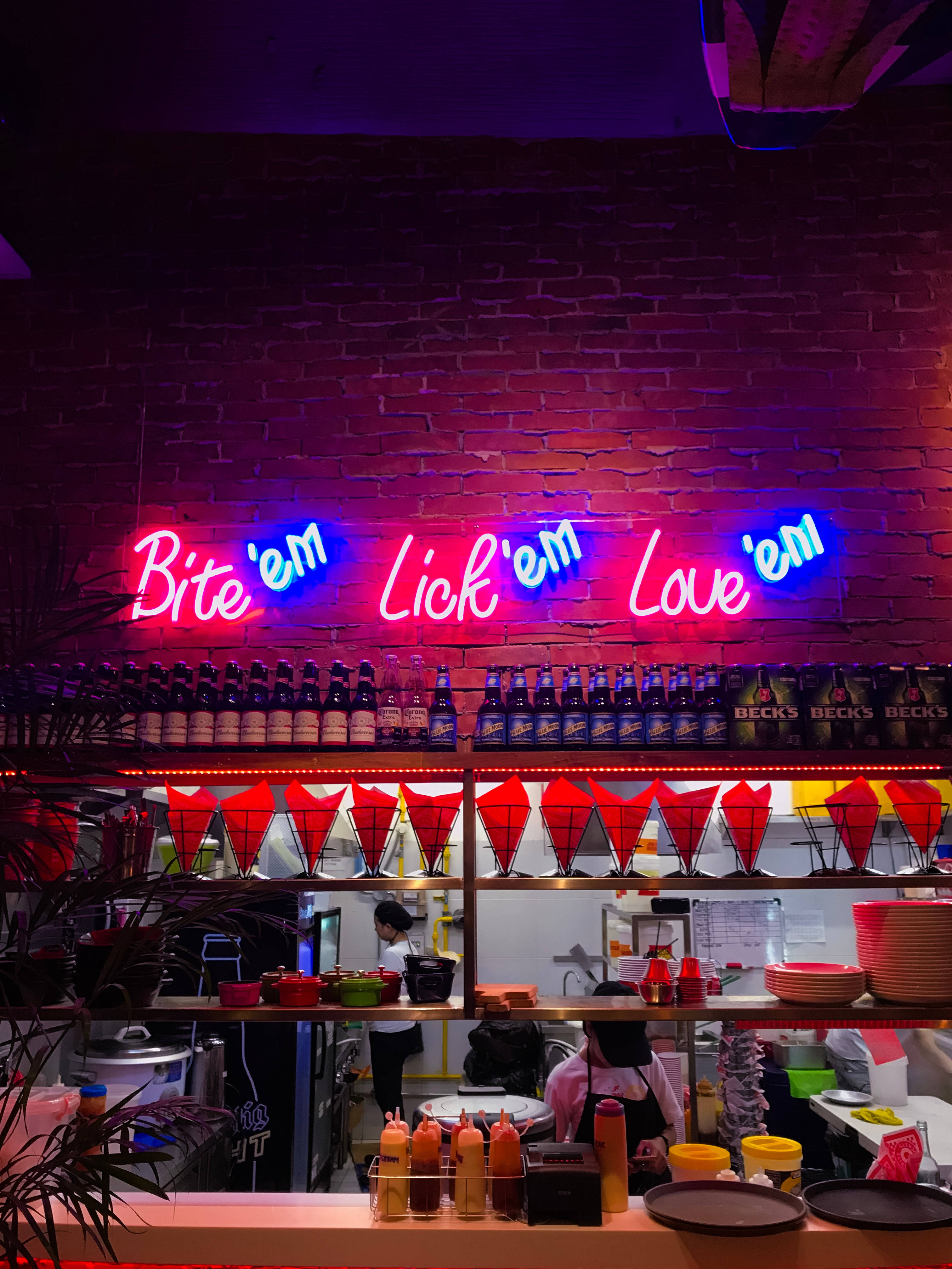 Neon Phone 桌布 壁紙 — 充滿活力的霓虹招牌，上面寫著 Bite'em Lick'em Love'em，在磚牆上閃爍著粉紅色和藍色光芒，位於繁忙的食物櫃檯上方。