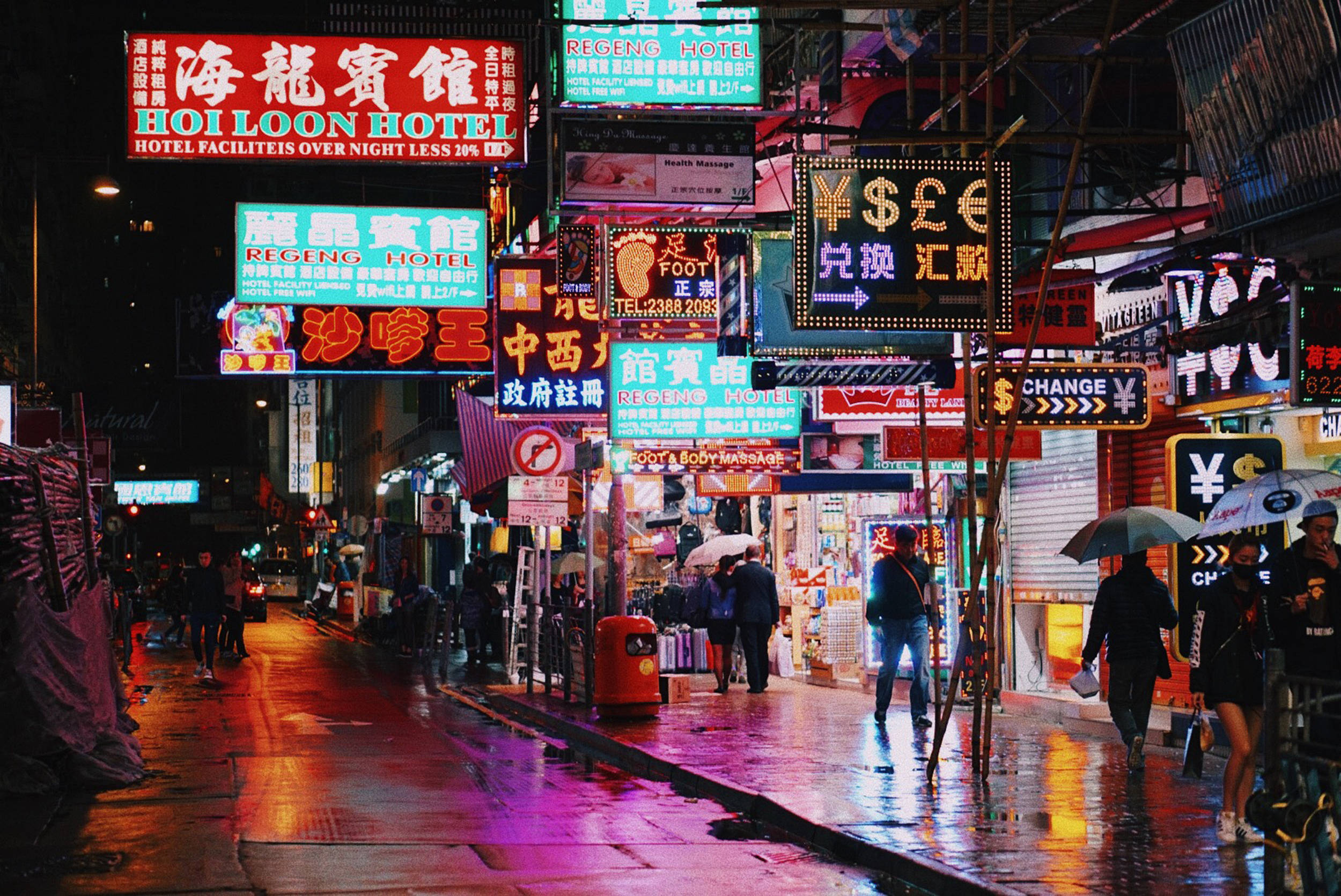 香港 桌布 壁紙 — 充滿活力的霓虹燈在雨濕的夜間城市街道上閃耀，行人撐著傘在濕滑的人行道上行走。