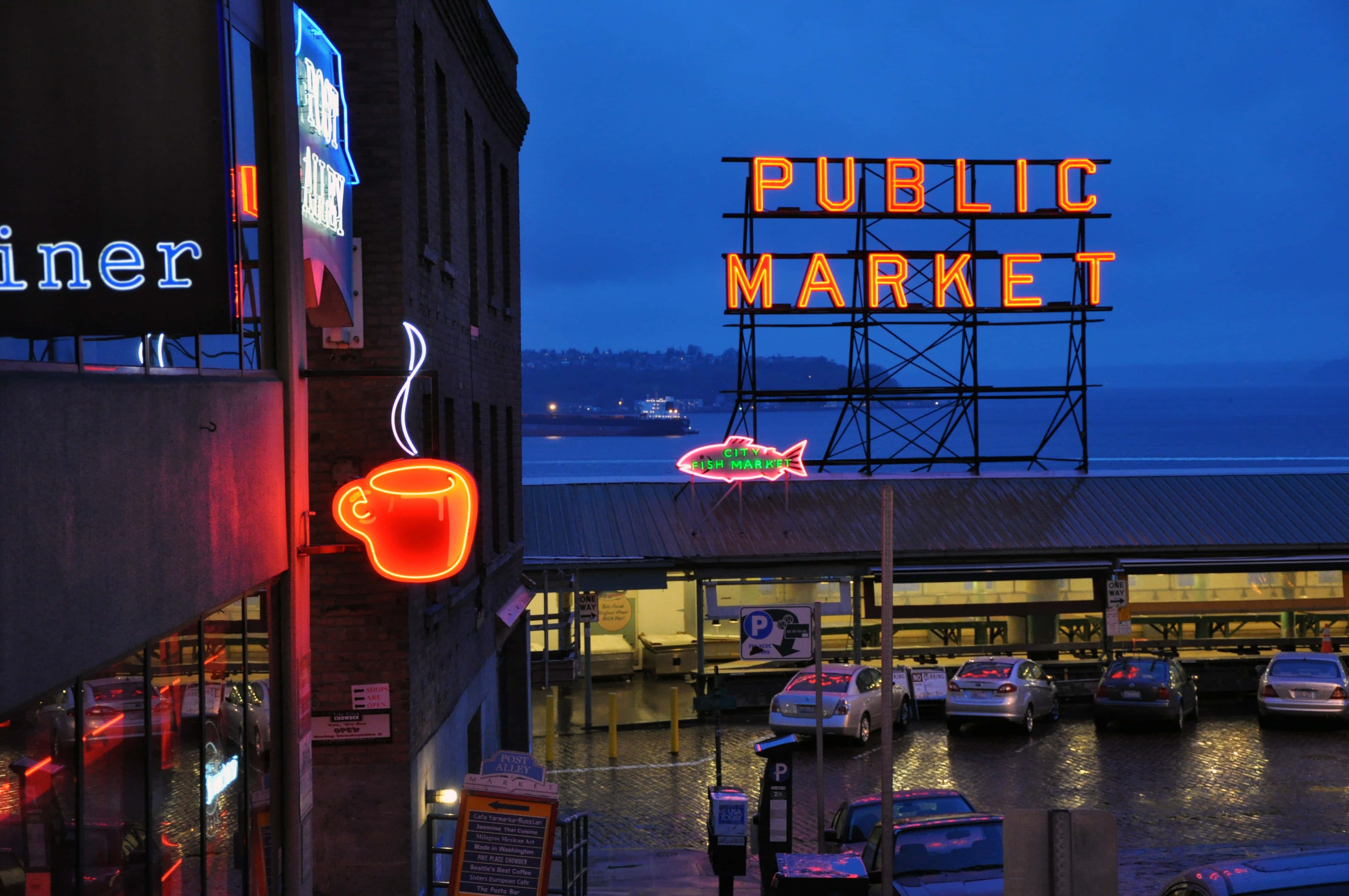 Seattle At Night 桌布 壁紙 — 充滿活力的霓虹燈在黃昏時分照亮潮濕的城市市場，映照在雨水濕滑的路面上。