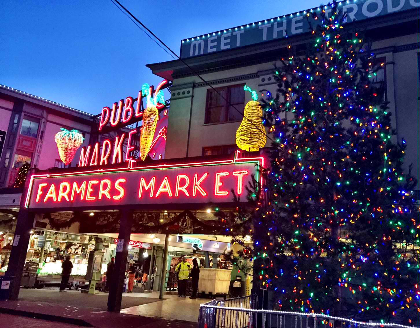 Pike Place Market 桌布 壁紙 — 充滿活力的霓虹燈招牌照亮了黃昏時分熙熙攘攘的農貿市場入口，旁邊是一棵璀璨奪目的聖誕樹。