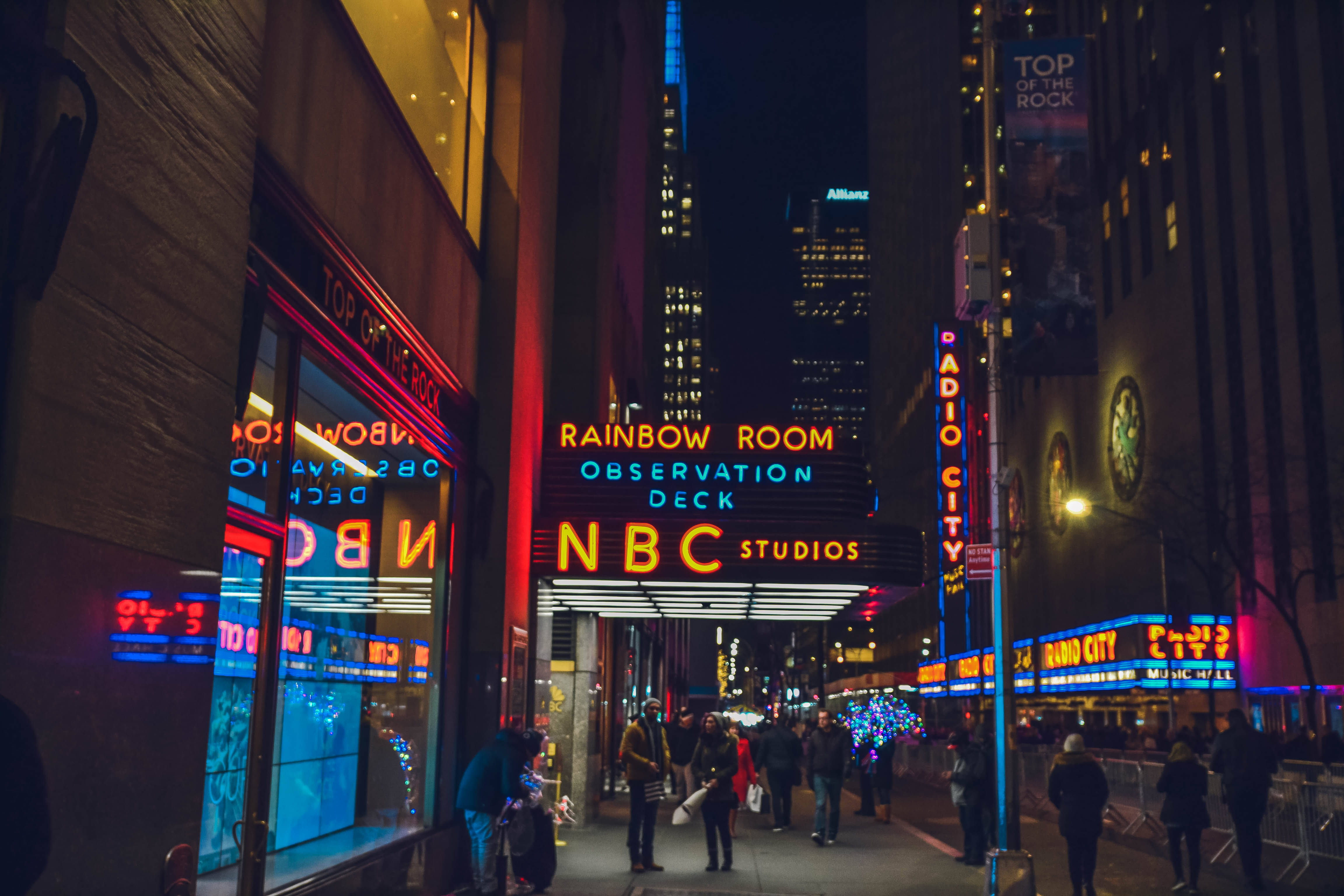 Nyc 桌布 壁紙 — 充滿活力的霓虹燈照亮了夜晚繁忙的紐約市街道，行人走過標誌性的NBC和無線電城大樓。