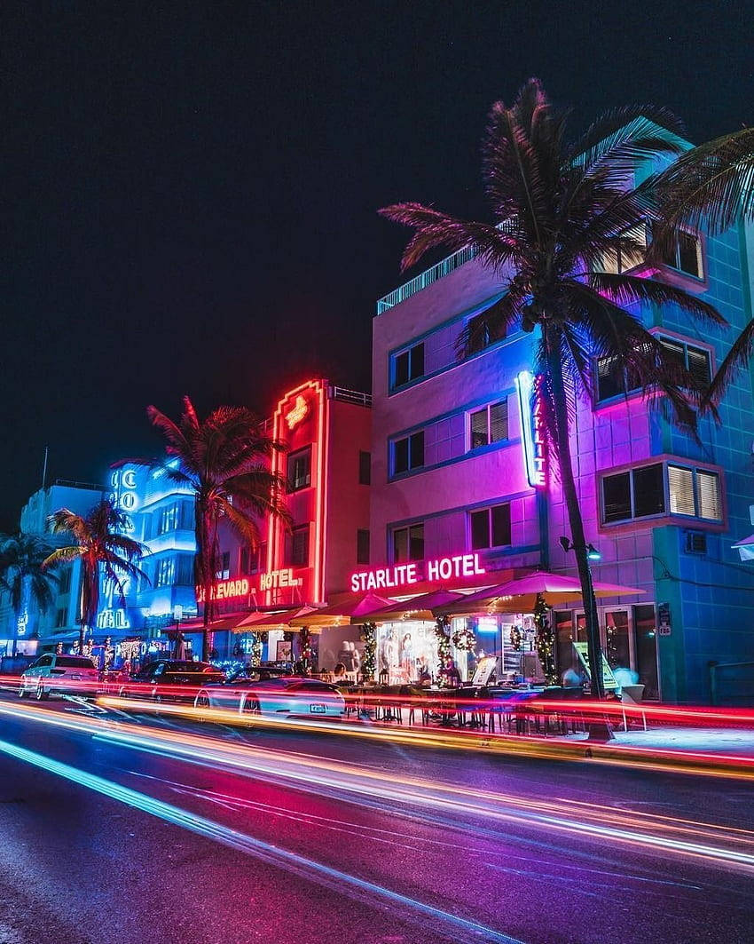 neon aesthetic iPhone 桌布 壁紙 — 充滿活力的霓虹燈照亮了裝飾藝術風格的酒店和棕櫚樹，汽車燈光形成的線條在邁阿密的夜晚街道上模糊地掠過。