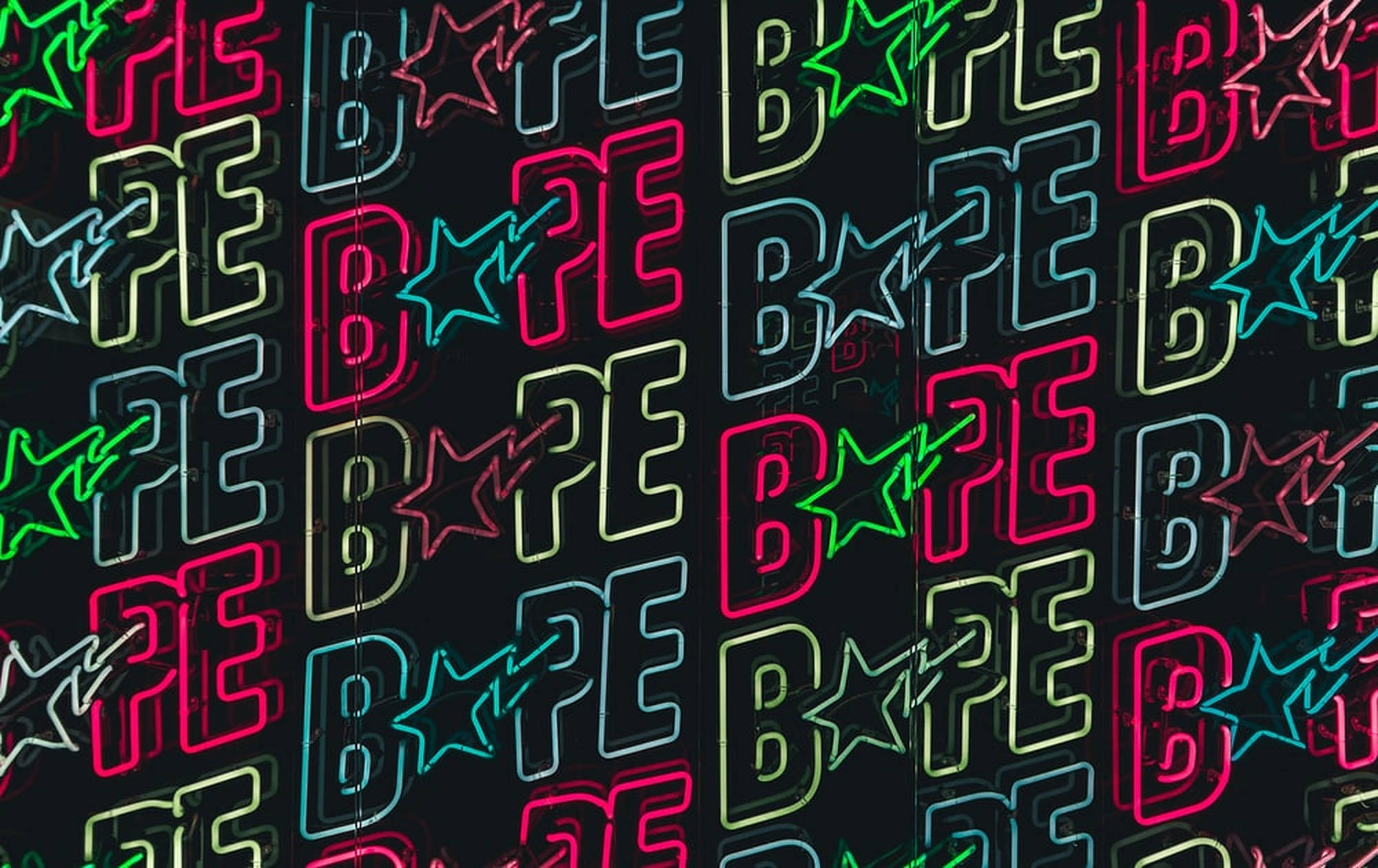 bape logo 桌布 壁紙 — 充滿活力的霓虹燈牌，重複的「BAPE」標誌和星星在粉紅色、綠色和藍色中發光，映襯著深色牆壁。