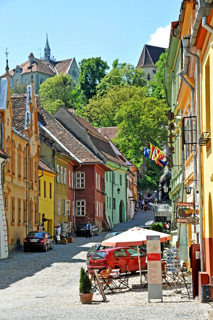 Romania 桌布 壁紙 — 充滿活力的鵝卵石街道，兩旁是色彩繽紛的歷史建築，在晴朗的藍天下，教堂尖塔聳立在茂密的綠樹之上。