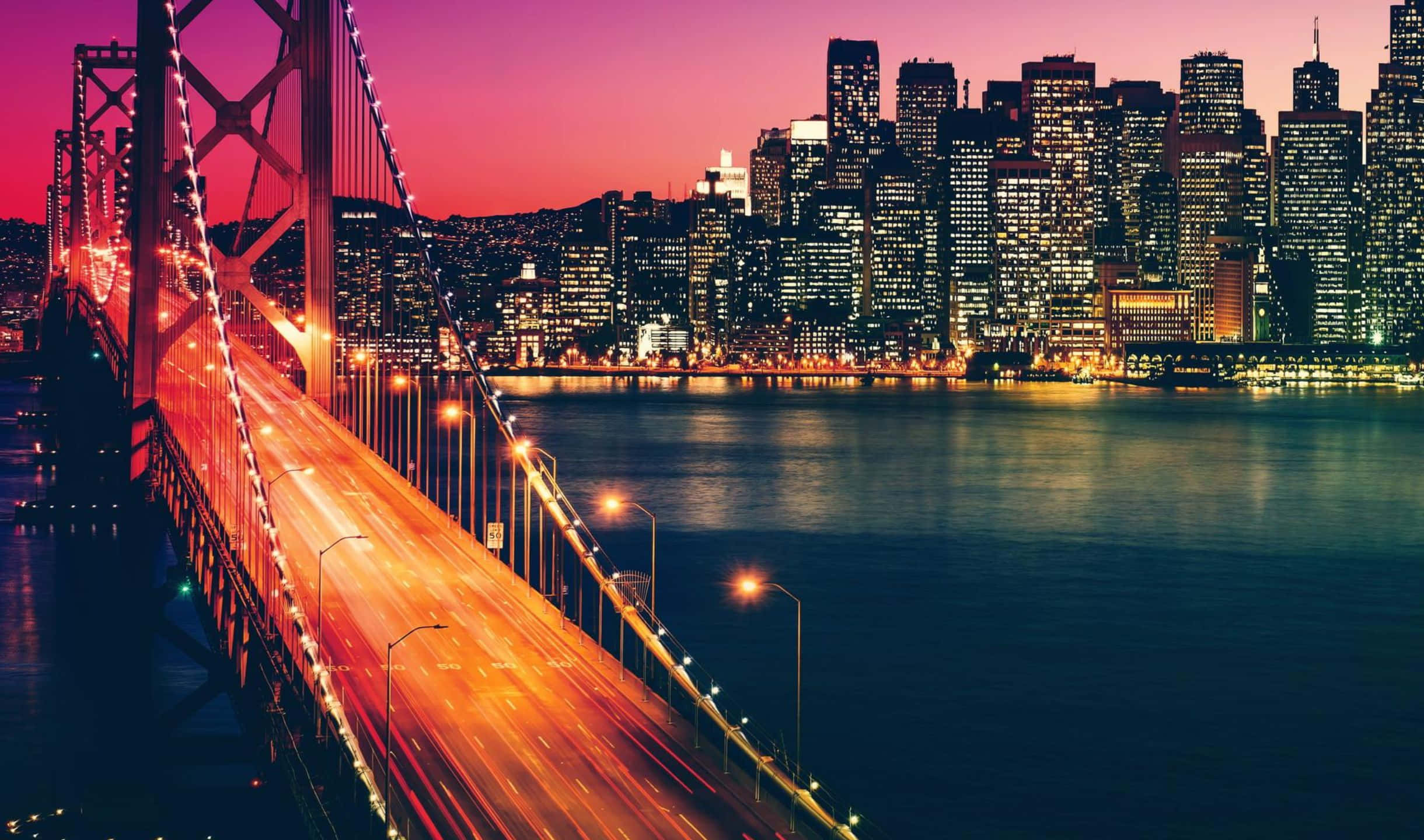 2440x1440 San Francisco 桌布 壁紙 — 充滿活力的黃昏城市景觀， featuring 一座發光的橋樑，水面上有流動的車燈軌跡，周圍是燈火通明的摩天大樓。