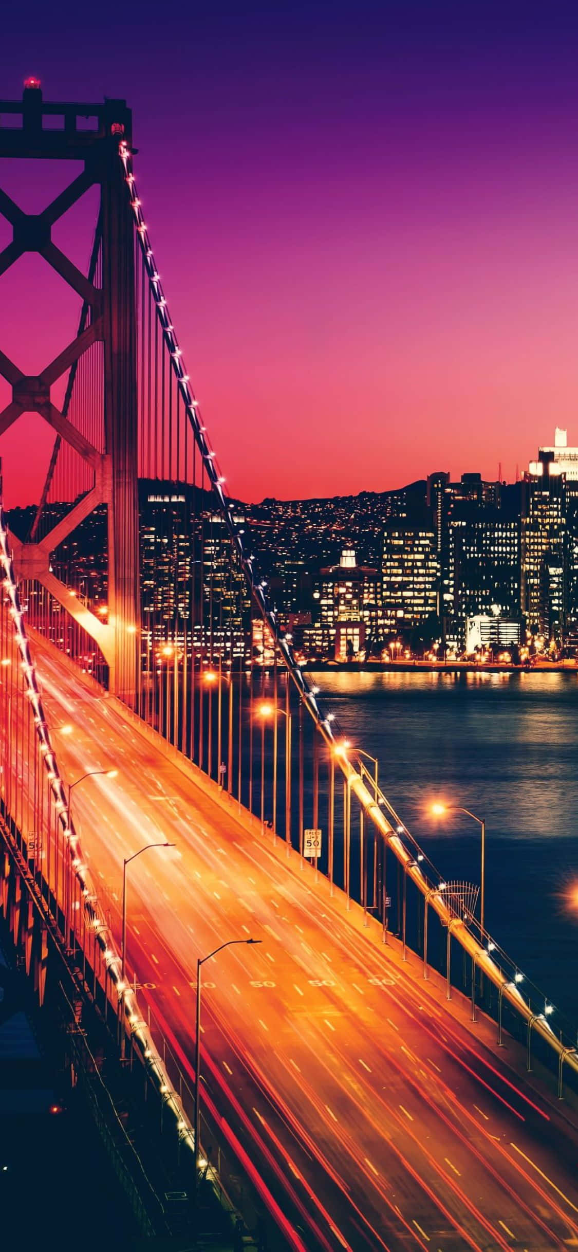 iPhone Xs California 桌布 壁紙 — 充滿活力的黃昏城市景觀，一座發光的吊橋，水面上有流動的交通燈光，以及燈火通明的摩天大樓。