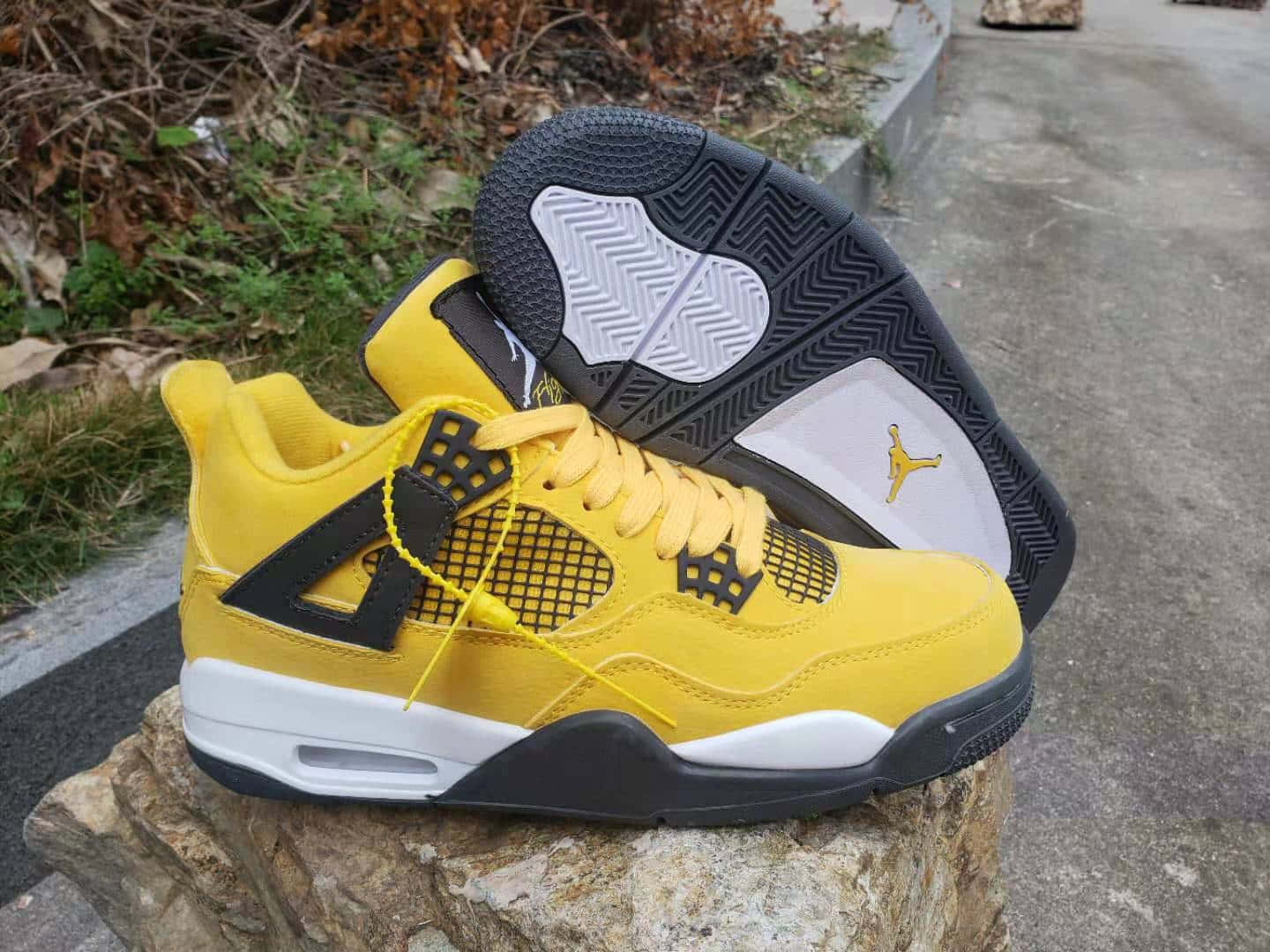 鞋子 桌布 壁紙 — 充滿活力的黃色 Jordan 4 運動鞋，帶有黑色點綴和白色鞋底，放置在風化的石頭上，背景是乾草和路面。
