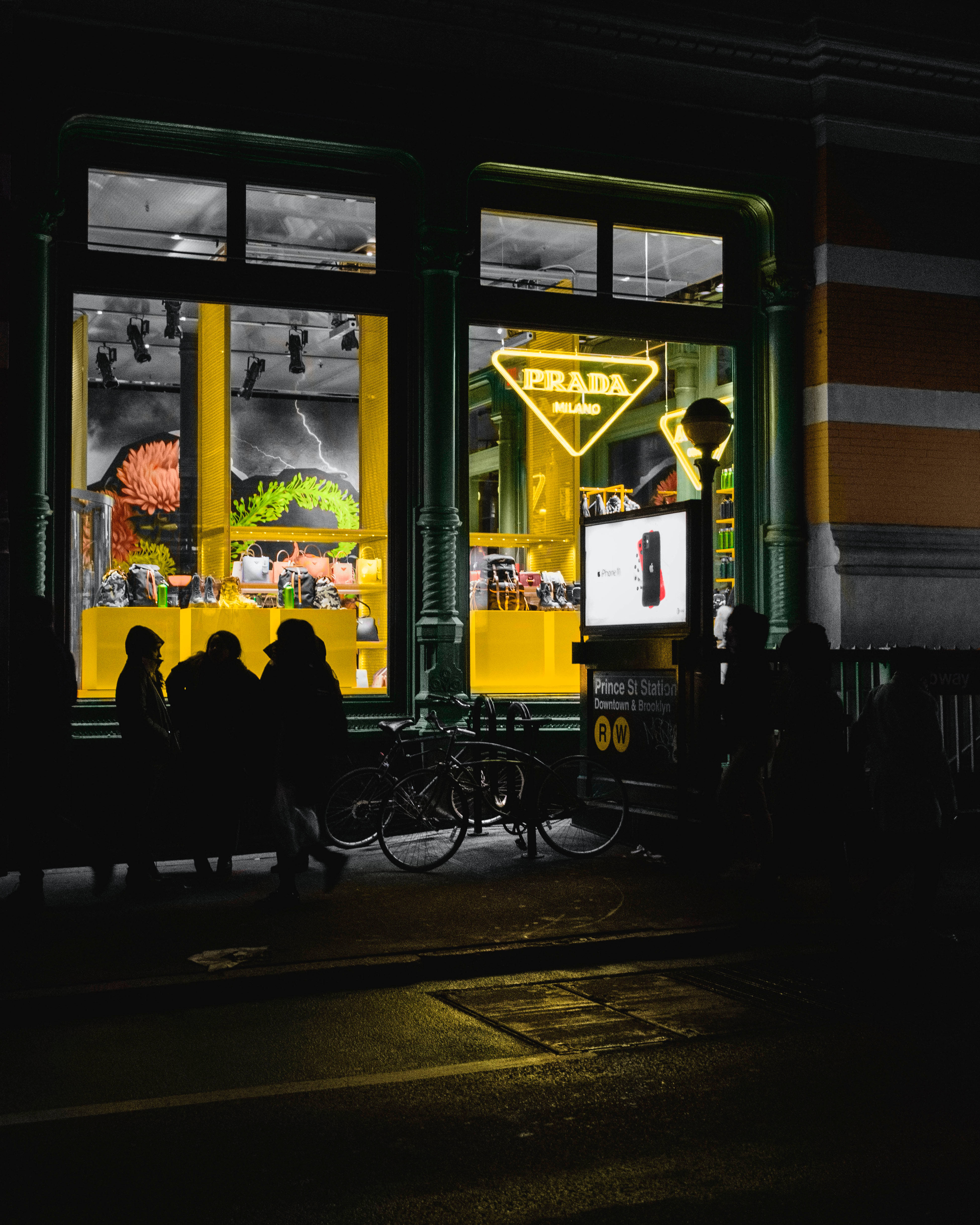 Prada 桌布 壁紙 — 充滿活力的黃色 Prada 商店櫥窗在夜晚發光，照亮黑暗城市人行道上的人和自行車的剪影。