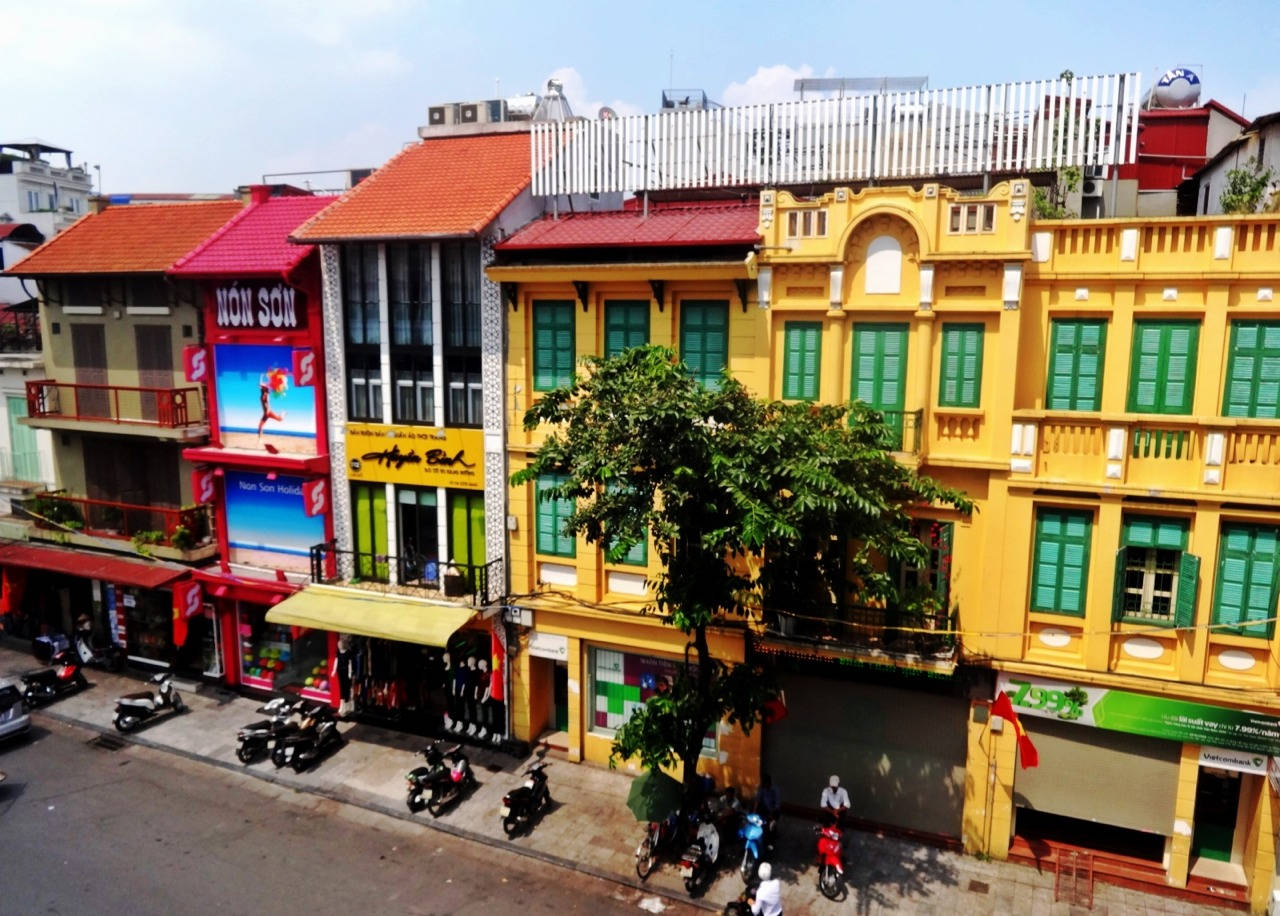 Hanoi 桌布 壁紙 — 充滿活力的黃色和紅色殖民建築排列在一條繁忙的街道上，停著摩托車和綠色百葉窗，在晴朗的藍天下。